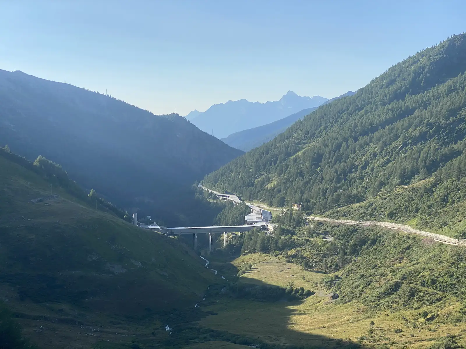 A forested valley descends toward distant snow-flecked peaks, a road bridge and stone retaining wall carrying the route across the hillside.