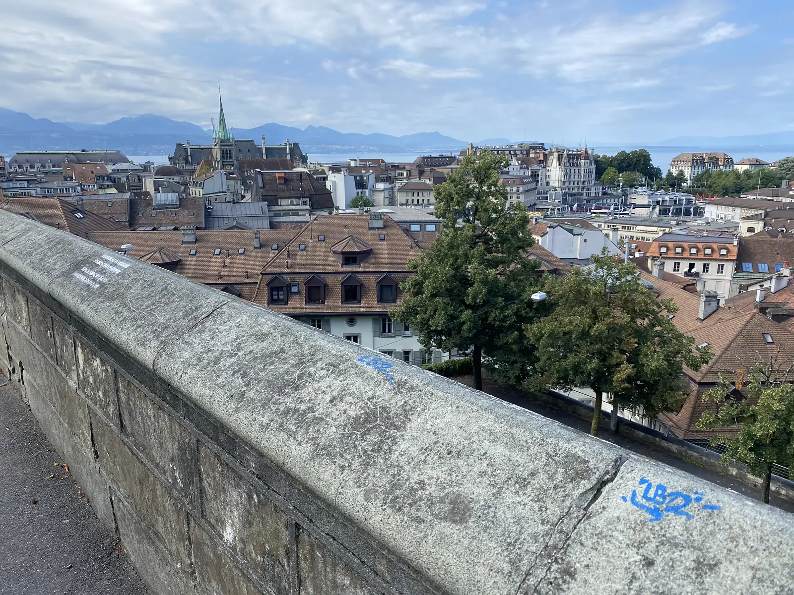A rooftop view over Lausanne's terracotta roofs and church spires towards Lake Geneva and the distant Alps, seen past a stone parapet marked with blue graffiti.