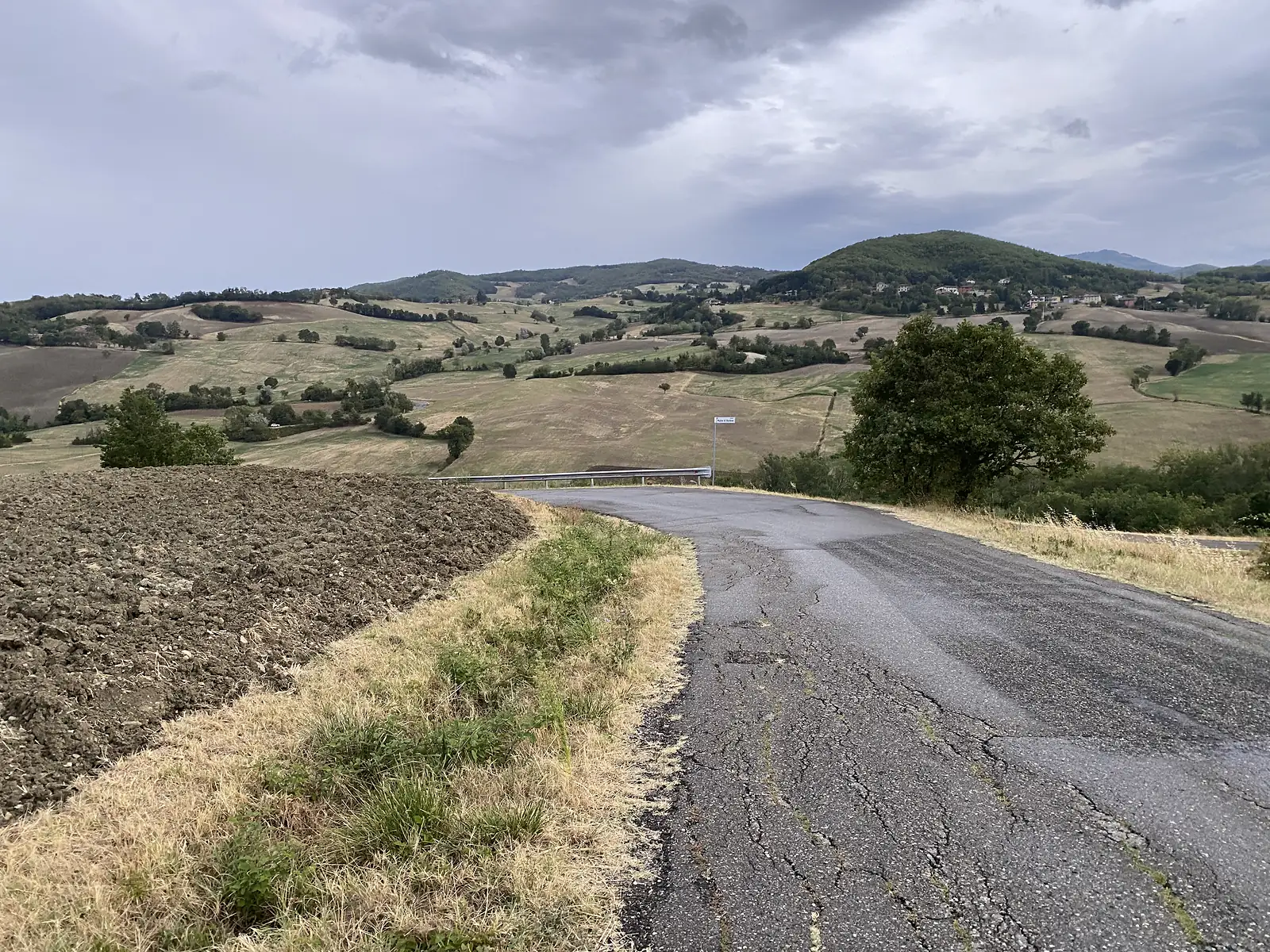 A cracked tarmac lane descends past a freshly ploughed field towards rolling Apennine hills under a heavy grey sky.