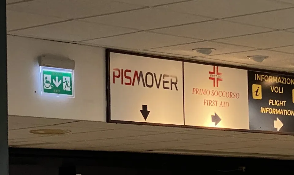 
An airport corridor sign reading 'PISMOVER', 'Primo Soccorso First Aid' and 'Informazioni Voli Flight Information', with an illuminated exit sign beside it.