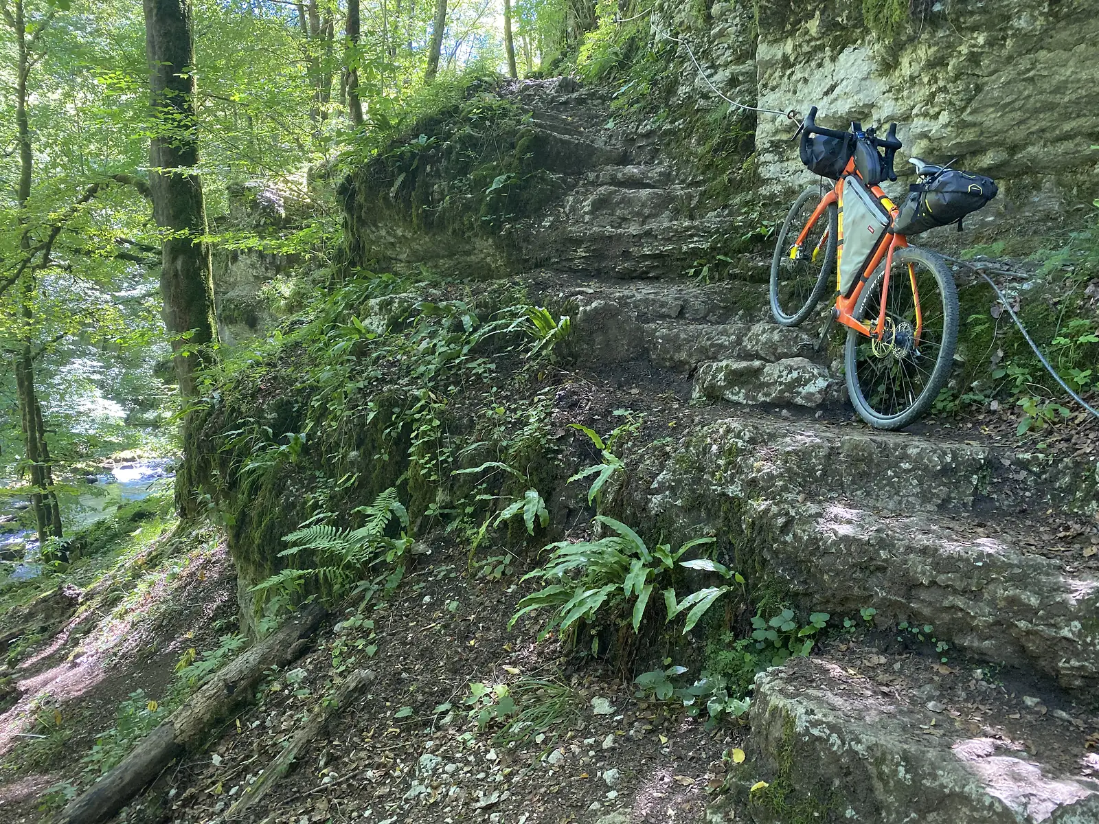 A loaded bike leans on a flight of rock-cut steps rising through a mossy cliff face above a gorge stream, a cable strung overhead.