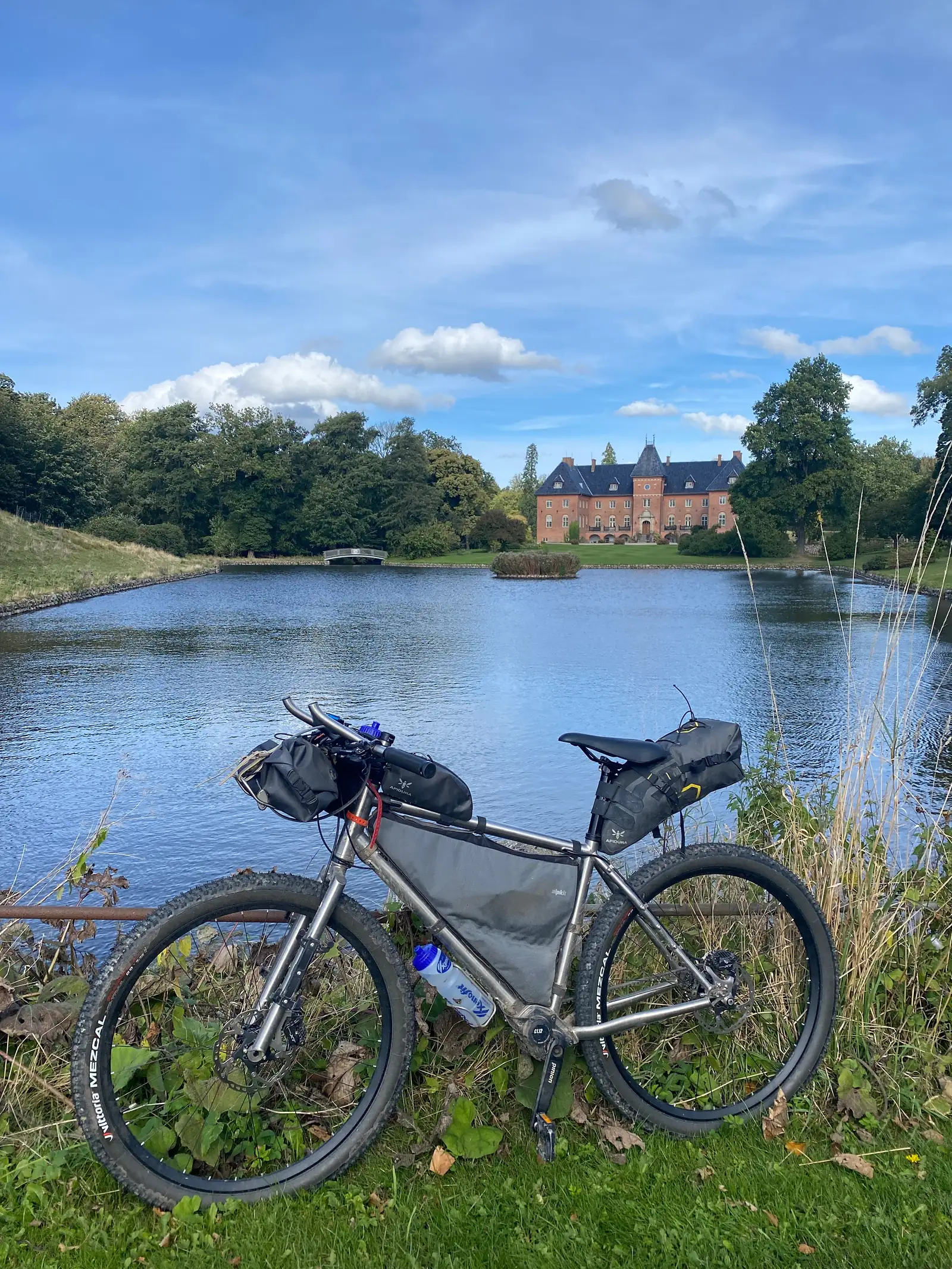 A loaded bikepacking bike propped on a rail beside a broad ornamental lake, with a red-brick manor and its bridge across the water.