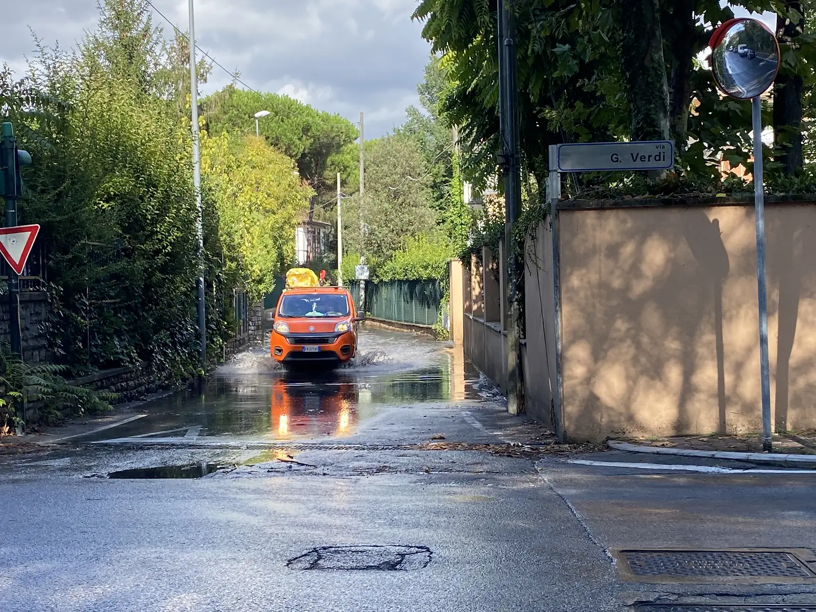 A small orange van splashes through a flooded dip in the road on Via G. Verdi, with a give-way sign and a traffic mirror at the corner in the foreground.