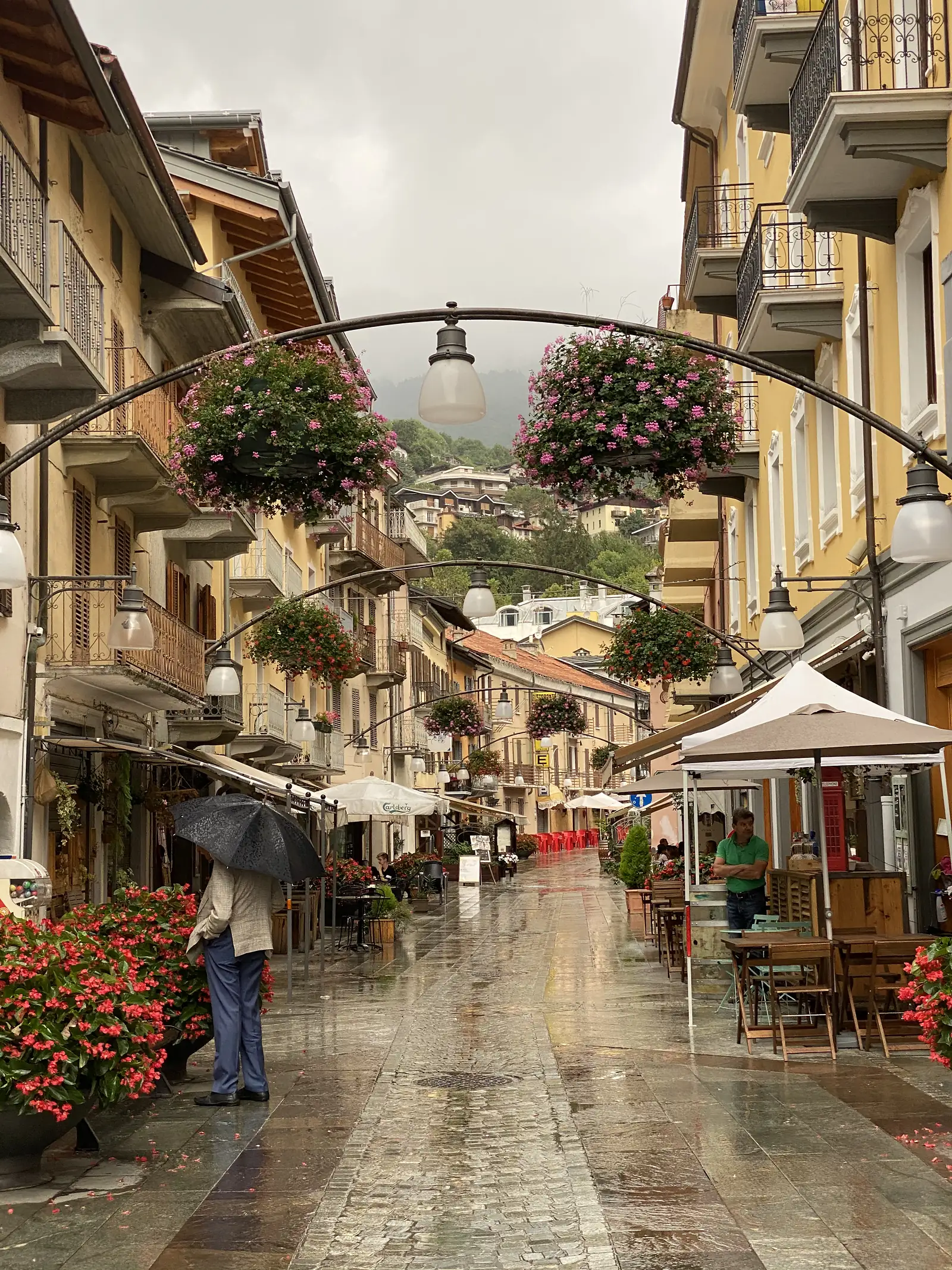 A rain-slicked pedestrian street in Saint-Vincent lined with yellow buildings, hanging baskets of pink geraniums strung over the road, and cafe tables under umbrellas.