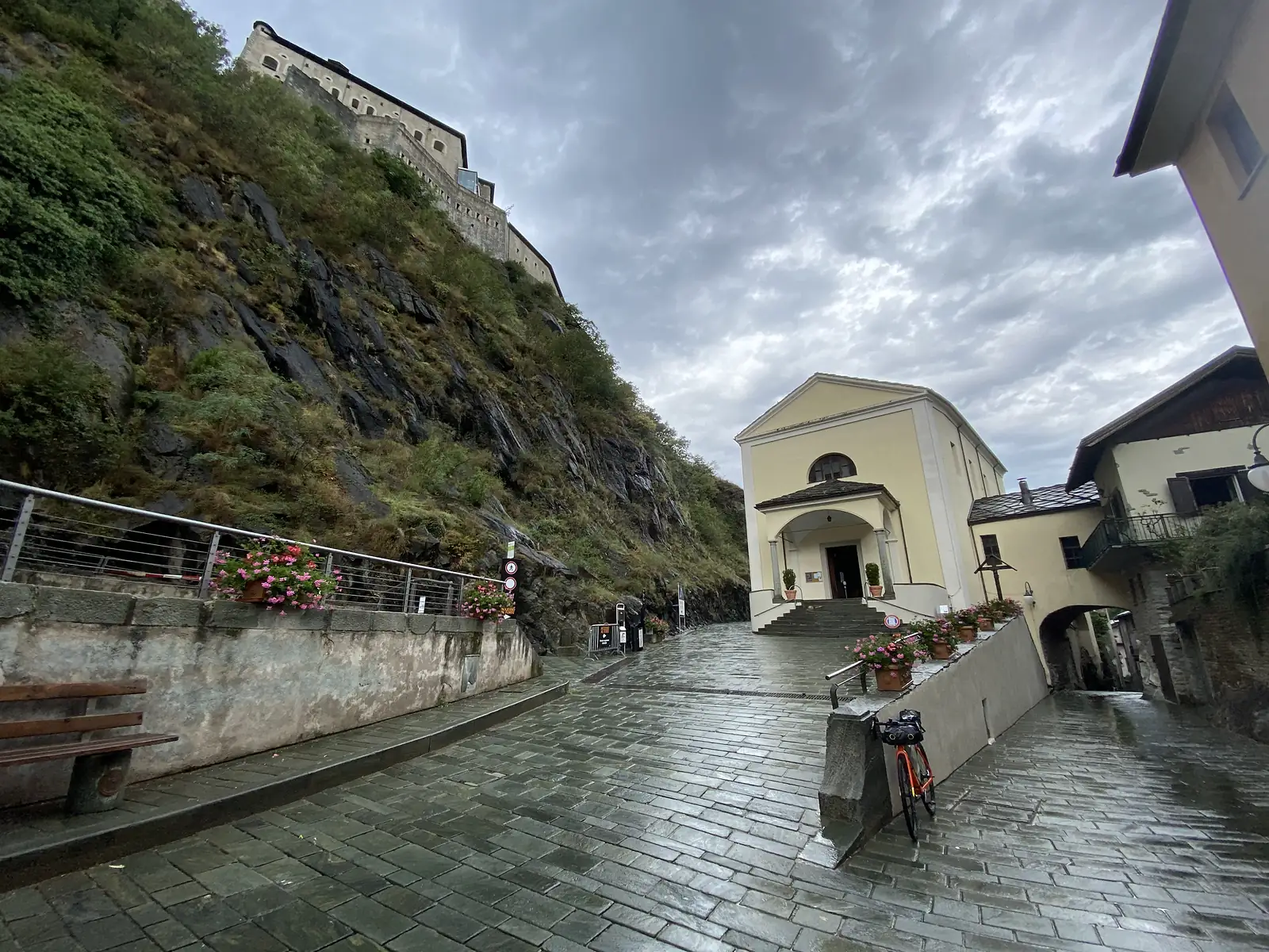 A loaded touring bike parked beside a small cream-coloured church at the foot of a cliff, with the Fort of Bard's ramparts visible on the rock above.