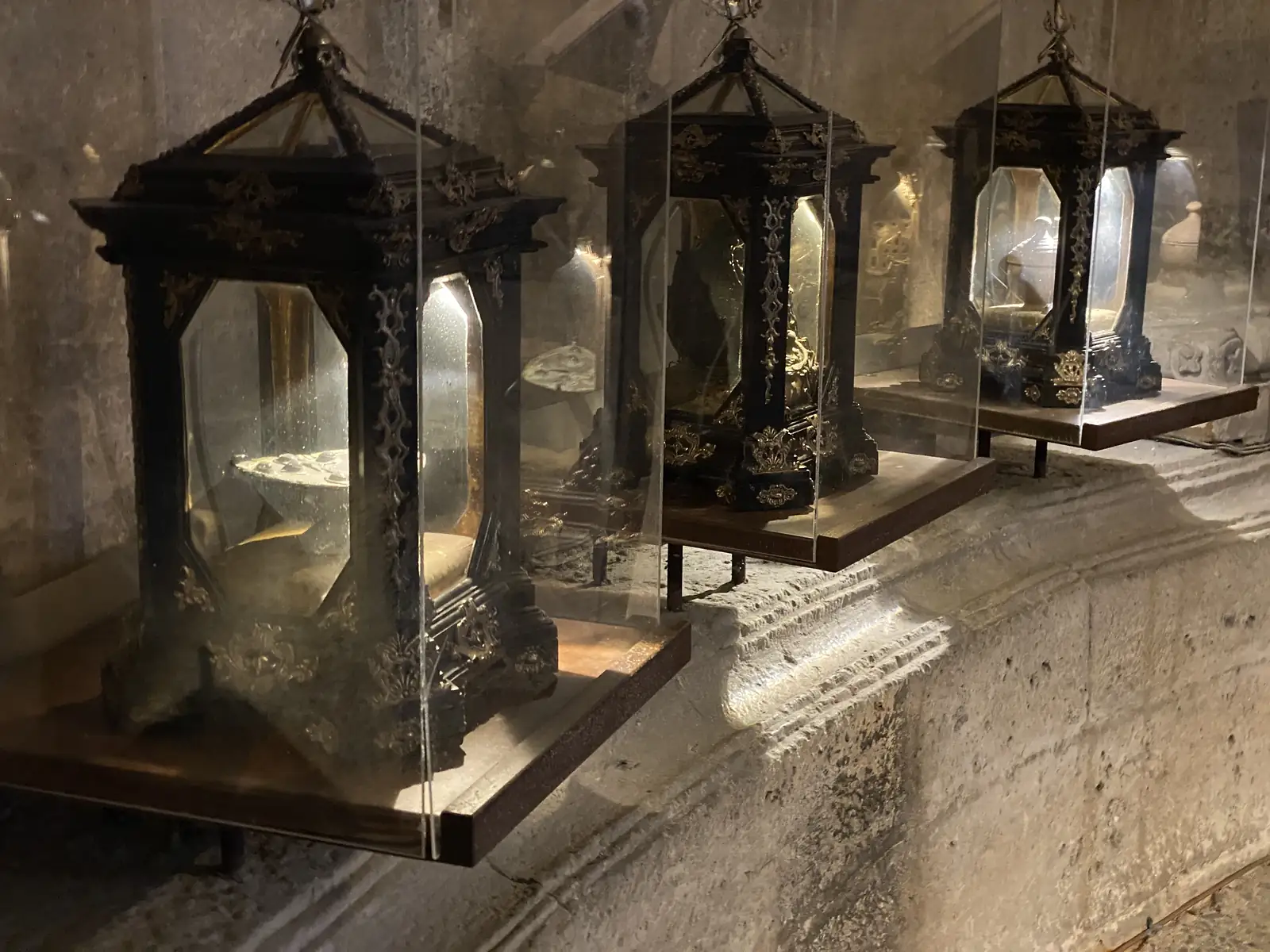A row of ornate reliquary shrines in dark wood and glass cases lines a stone wall inside a church treasury.