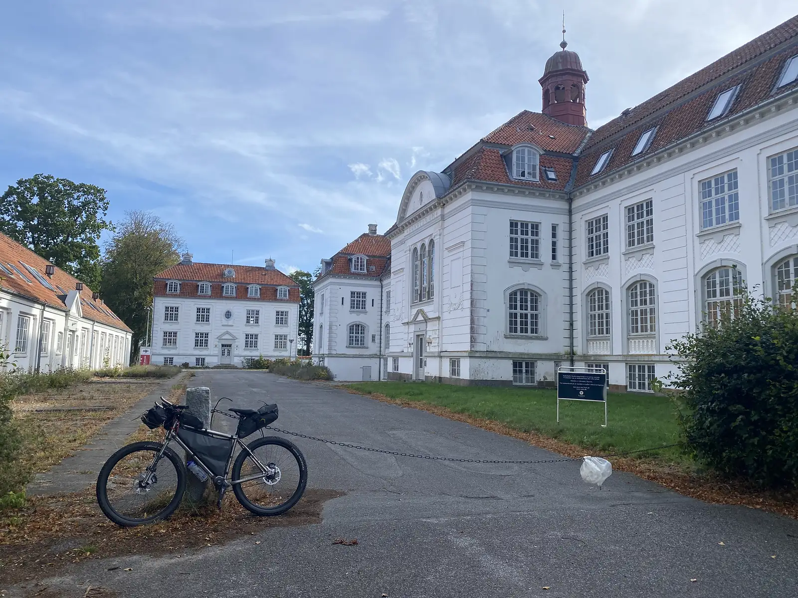 A loaded bikepacking bike leaning on a stone bollard on a weed-grown drive before a large empty white manor with a red bell turret.