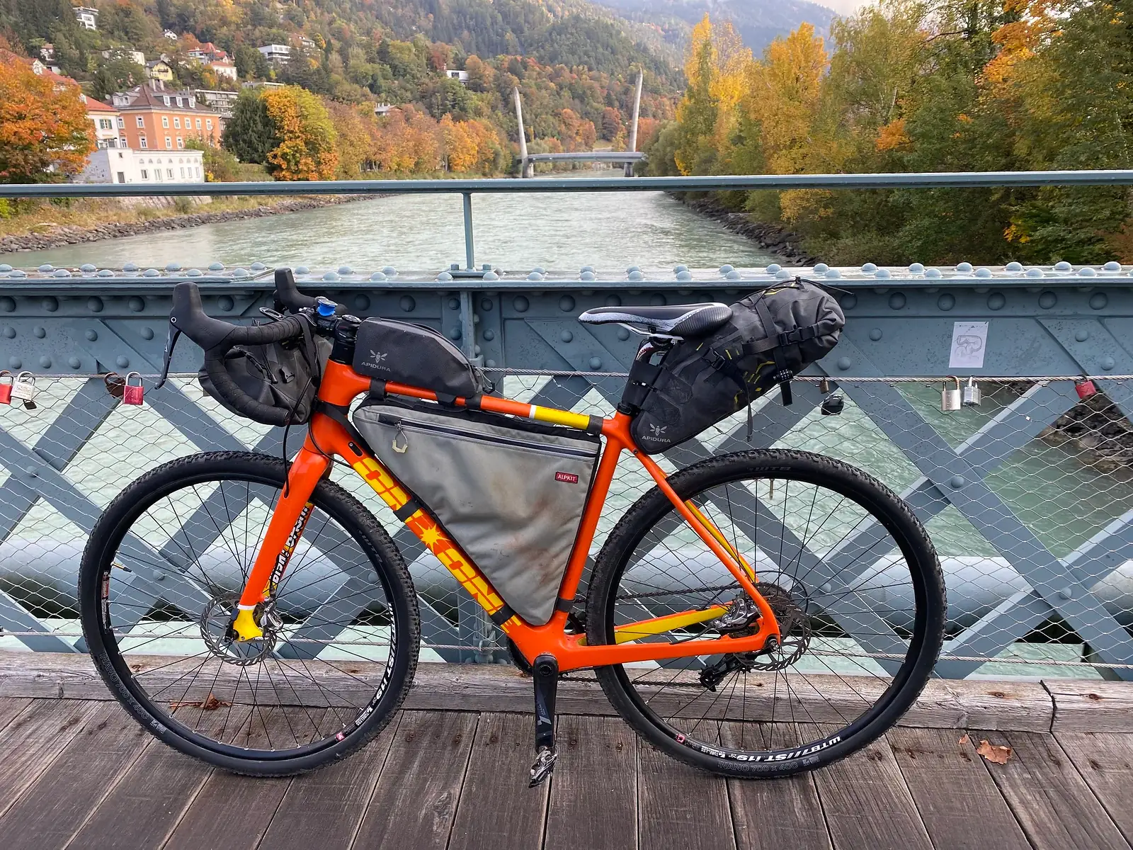 An orange On-One gravel bike loaded with bikepacking bags, propped against the railing of a river footbridge hung with padlocks, a hillside town and autumn woods behind.