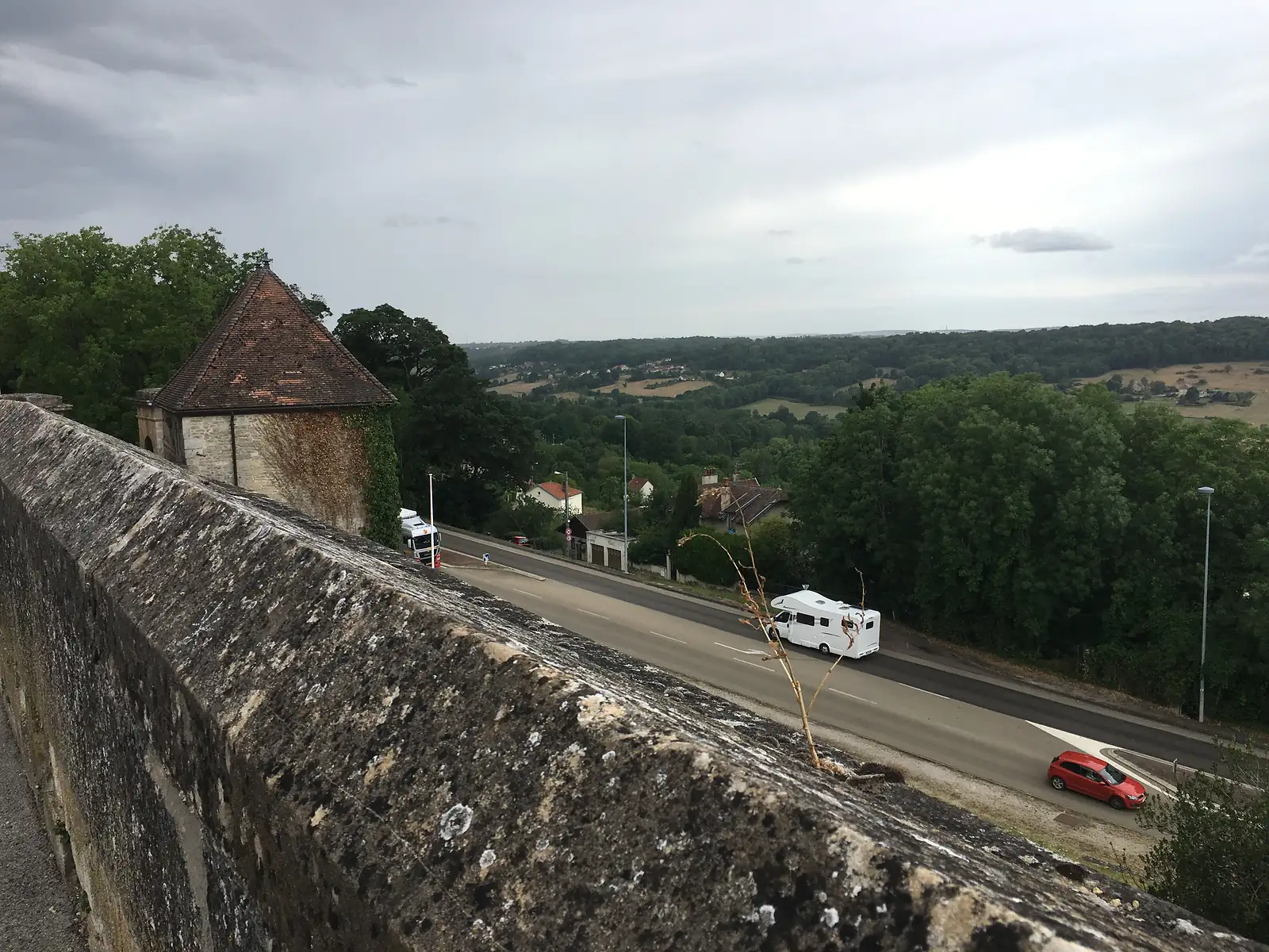 A view from the Langres ramparts over a road and a small stone tower roof, with fields and wooded countryside stretching to the horizon under a cloudy sky.