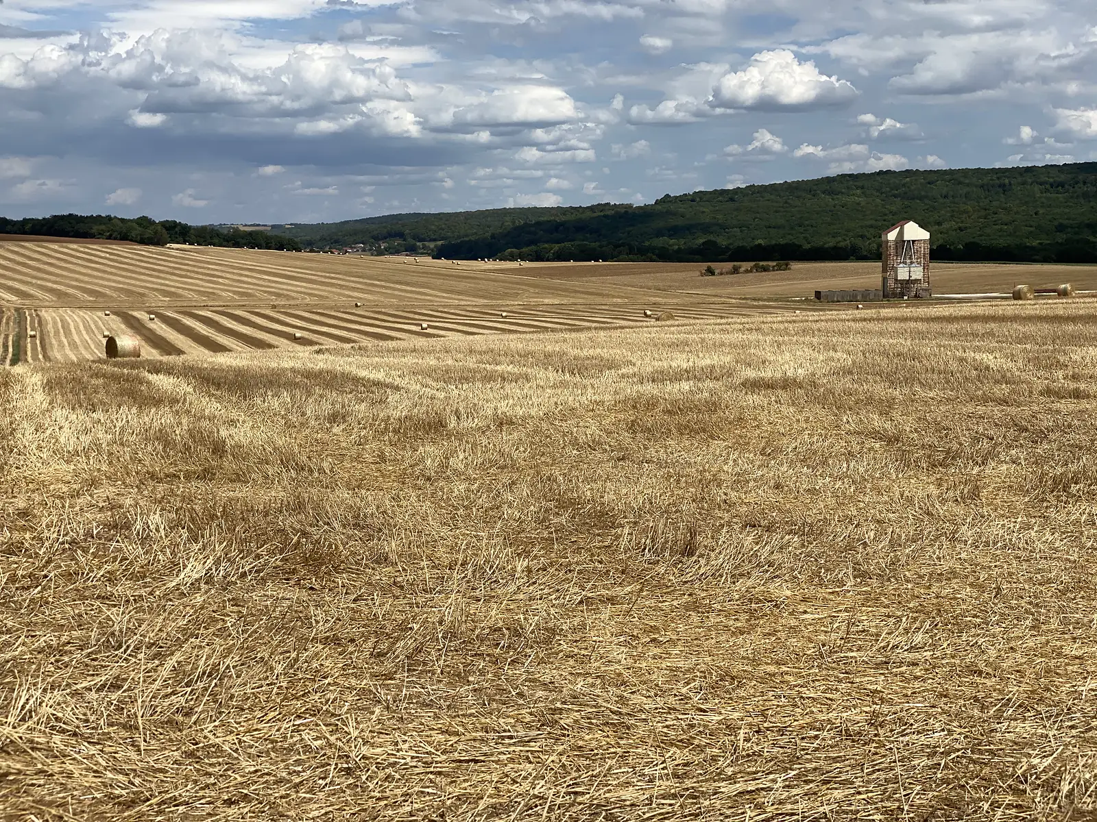 Rows of harvested stubble fields stretch toward a wooded ridge, a brick-and-white grain silo standing among scattered hay bales on the right.