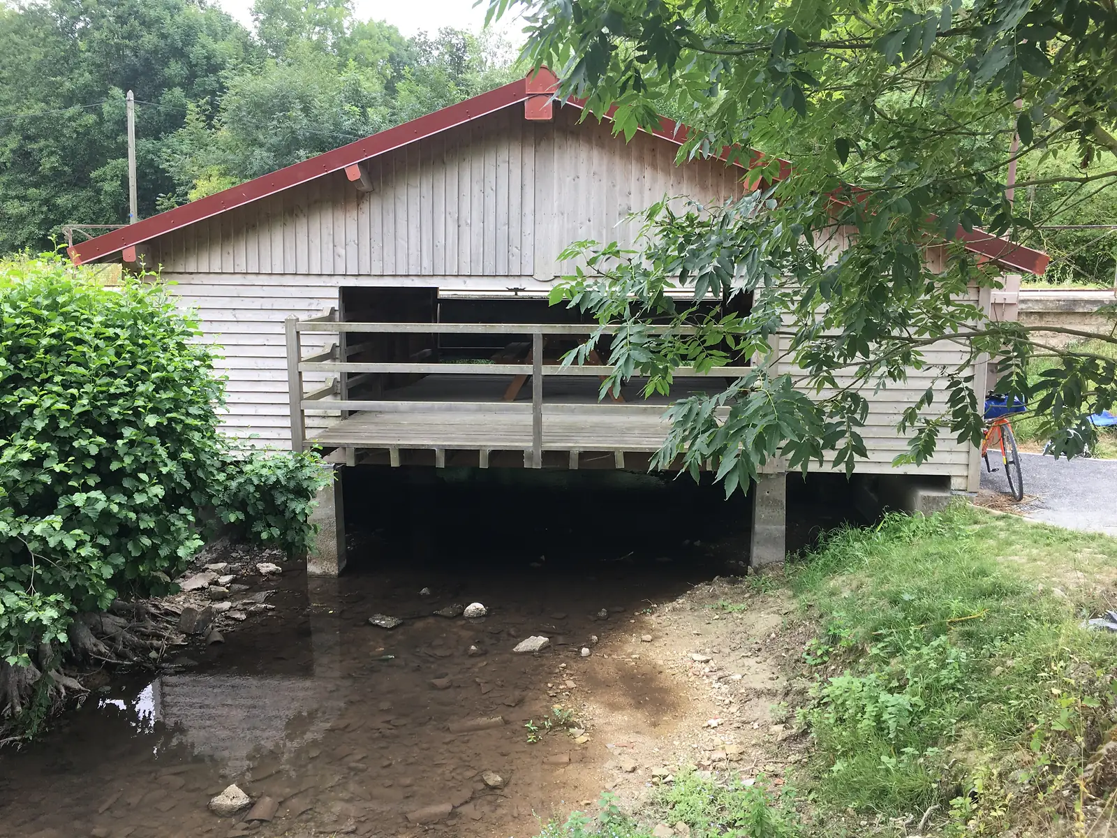 The timber-built shelter stands over a stream on concrete piers, its red ridge beam and pitched tiled roof visible through overhanging branches, with an orange bike parked by the doorway.