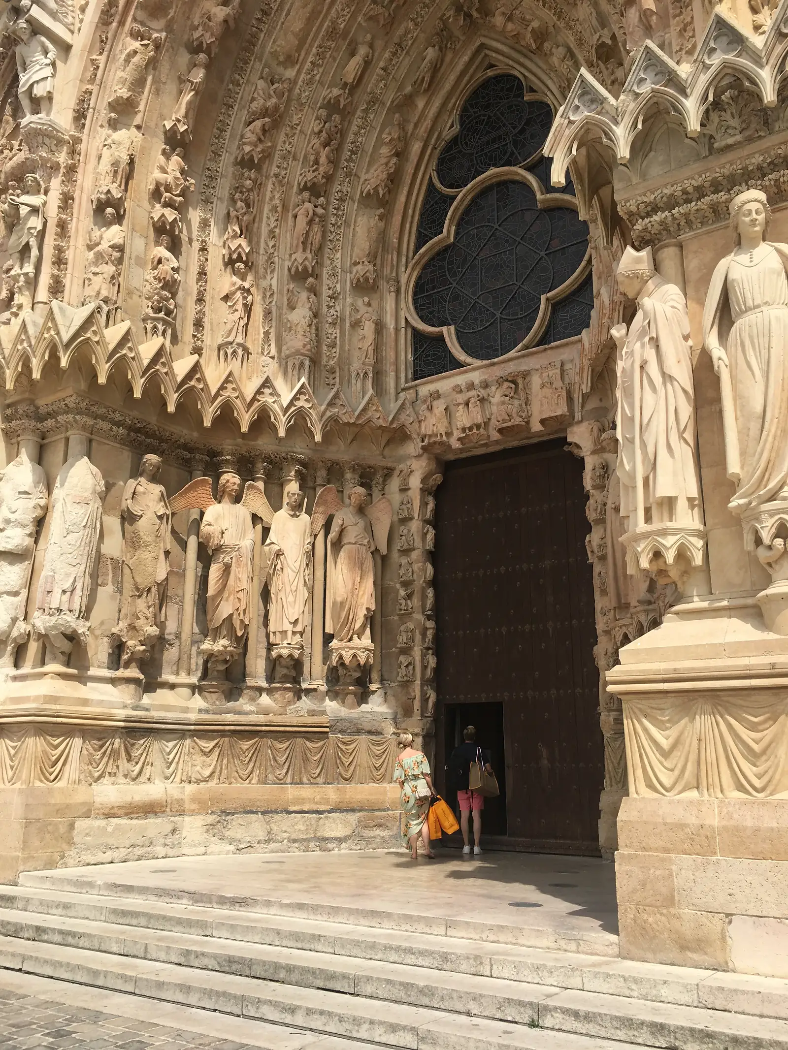 Weathered stone saints line a Gothic cathedral doorway above a quatrefoil window, with two visitors dwarfed beneath the huge dark wooden door.