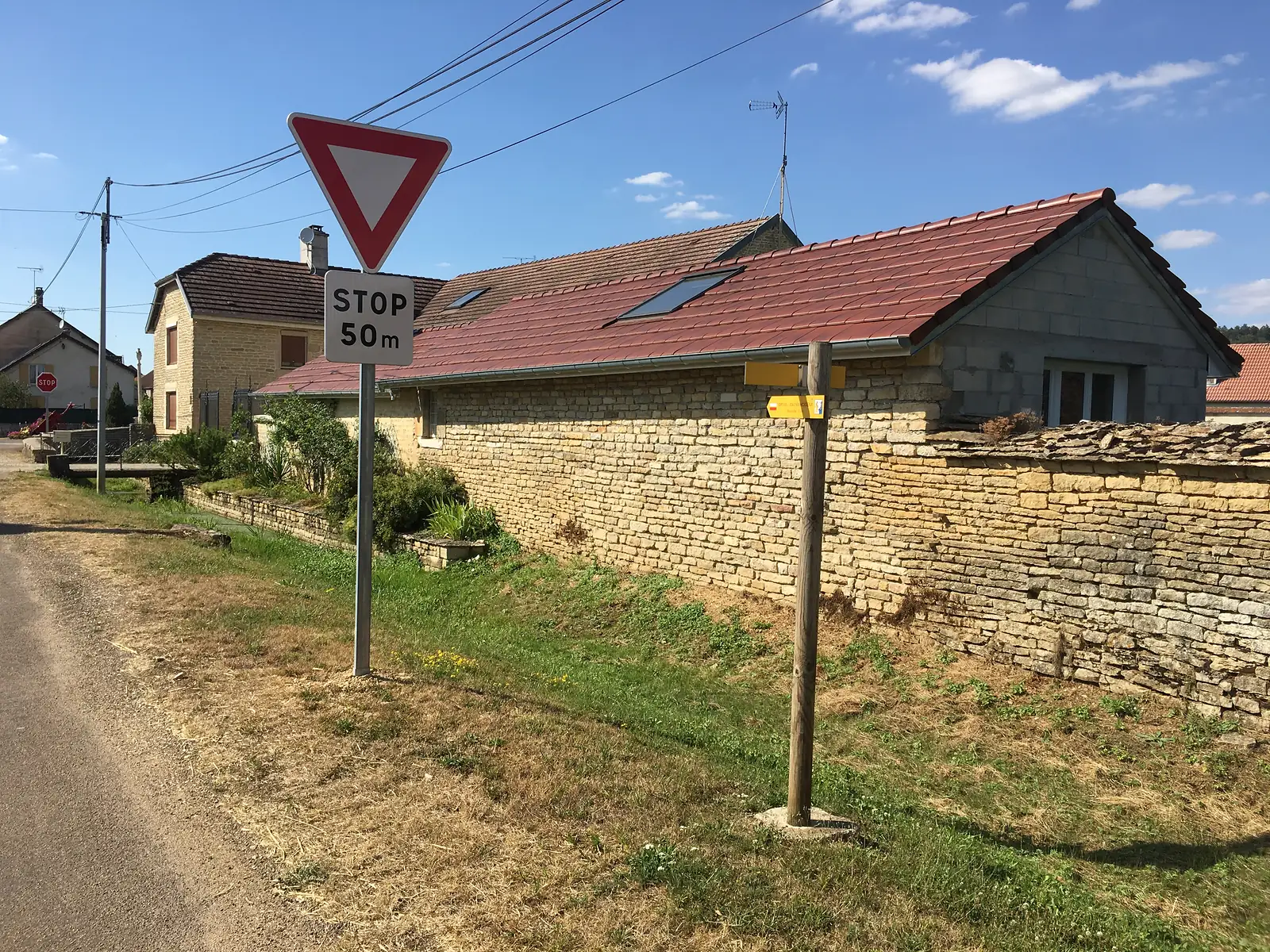A 'STOP 50m' sign and a yield sign stand beside a stone-walled house with a red tiled roof, with a small yellow Via Francigena marker on a post nearby.