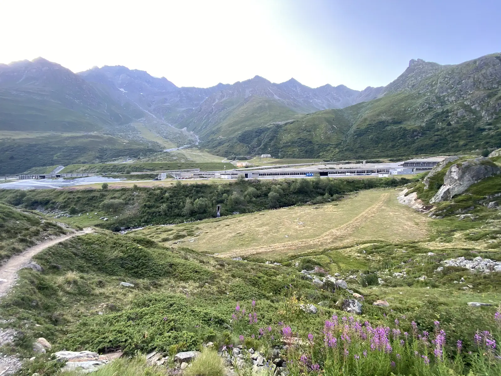 A broad mountain valley opens out around a cluster of low buildings beside a road, with peaks rising on both sides.