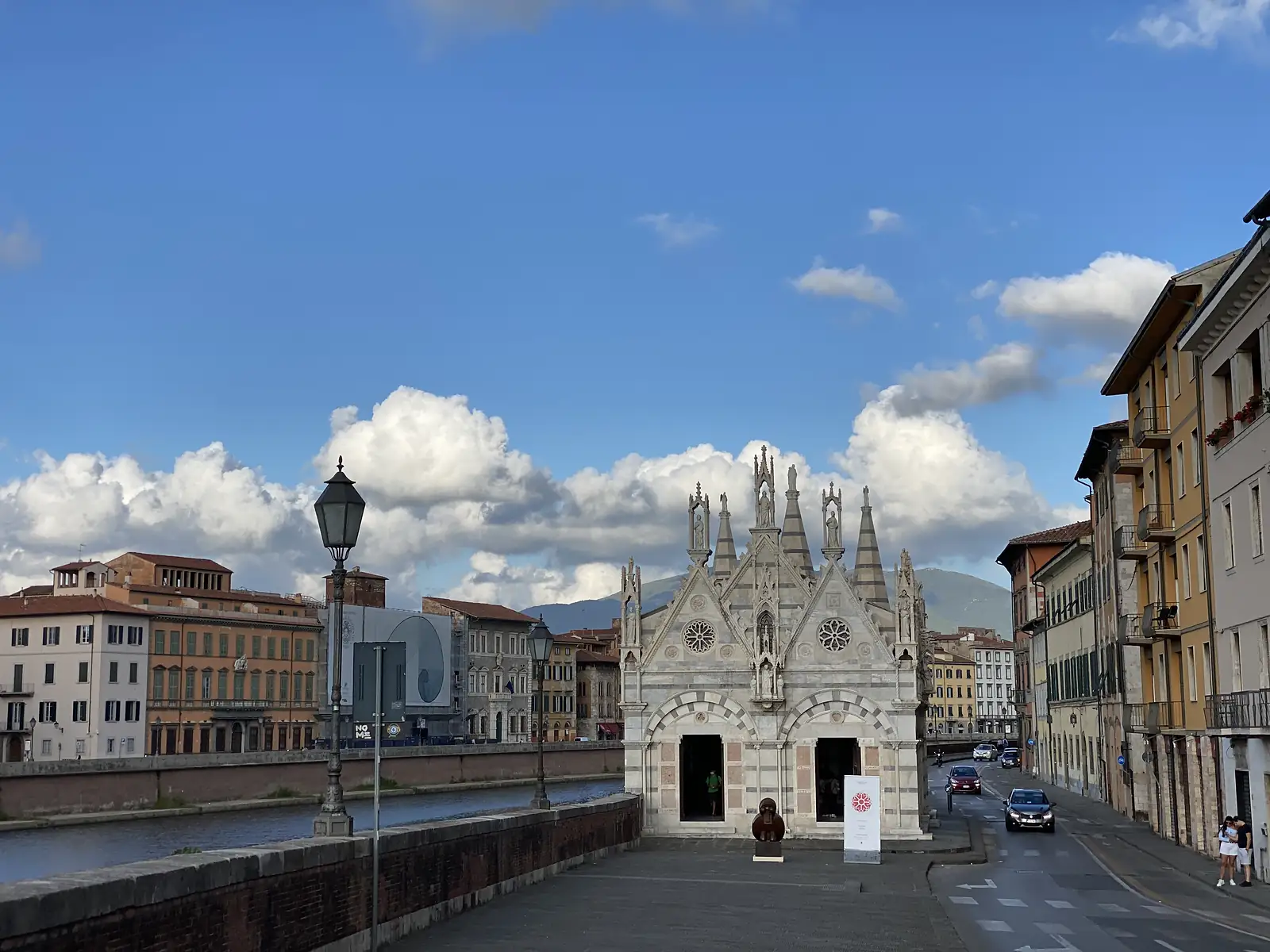 The small Gothic church of Santa Maria della Spina stands on the Arno embankment, its marble facade topped with spires and rose windows, beside a street lamp and passing traffic.