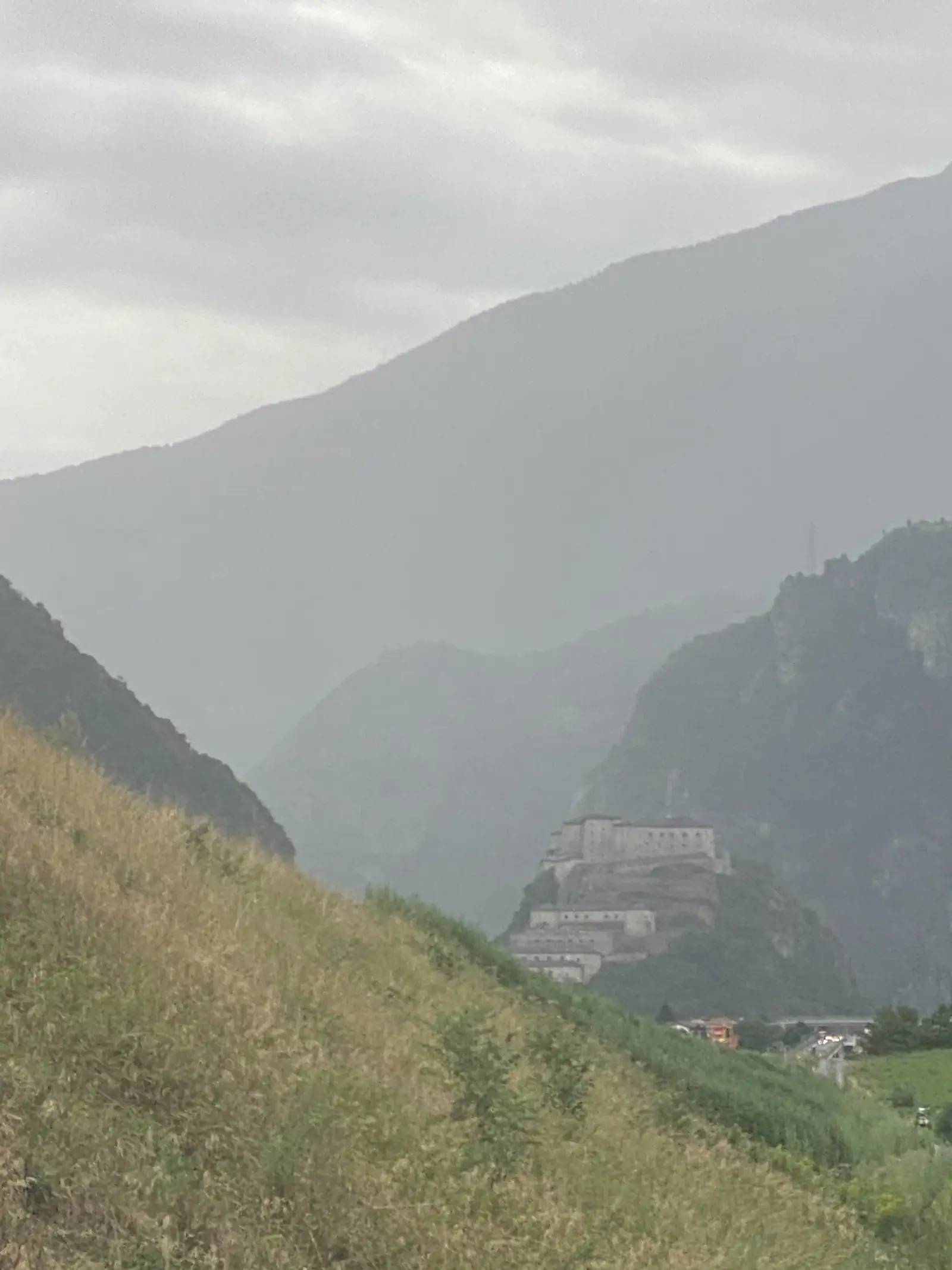 The tiered stone ramparts of a hilltop fort silhouetted against a misty mountain gorge, viewed from a distance across a valley.