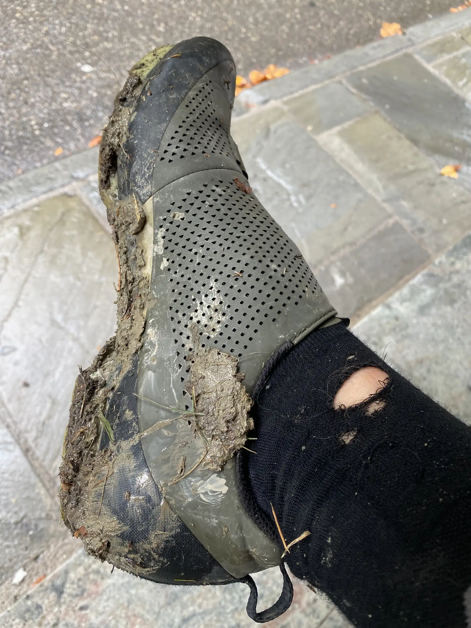A close-up of a mud-caked cycling shoe and torn black sock, worn through at the ankle, resting on a paved street.
