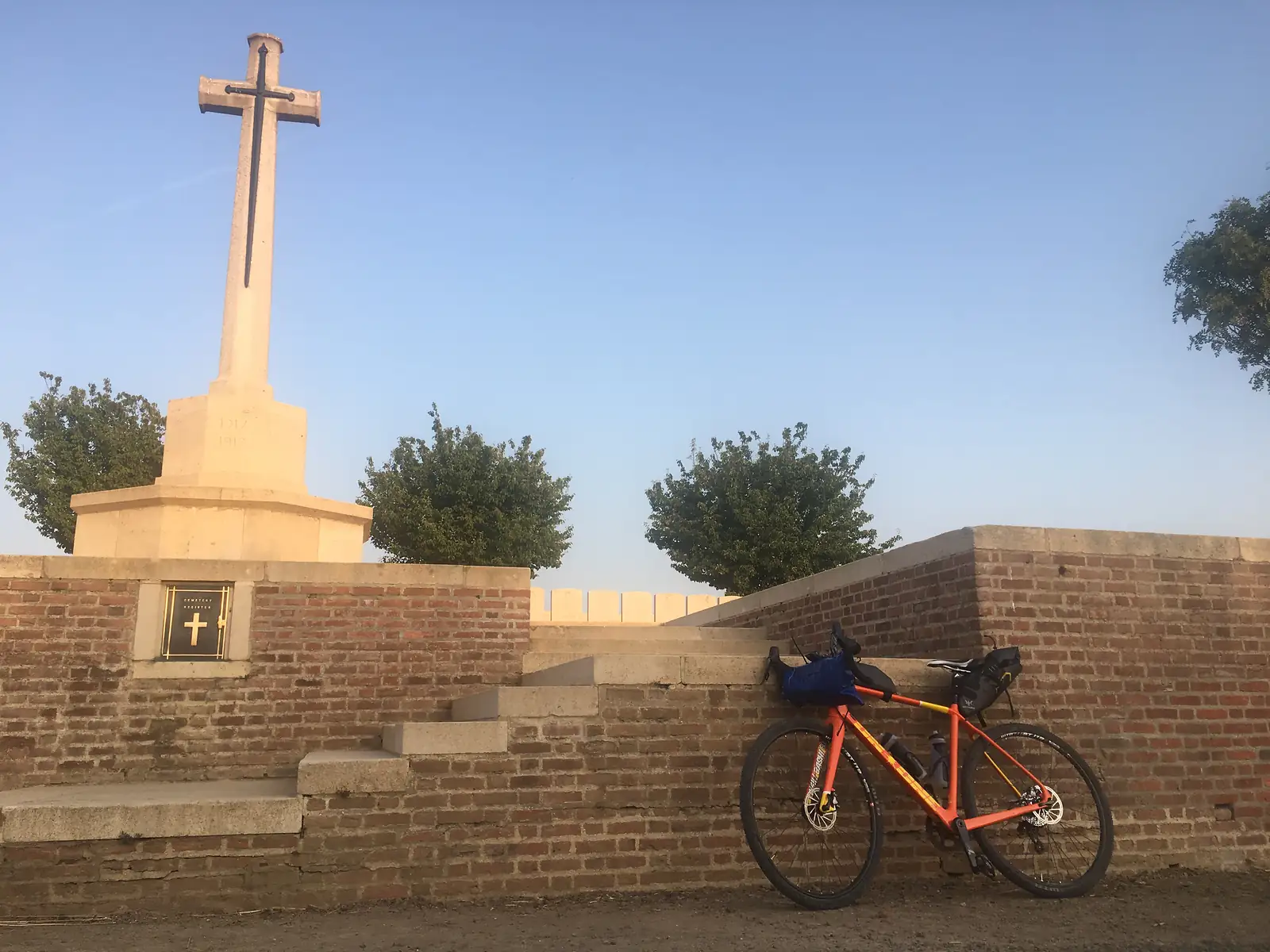A loaded orange gravel bike leaning against the brick boundary wall of a war cemetery, its Cross of Sacrifice rising behind steps and rows of headstones.