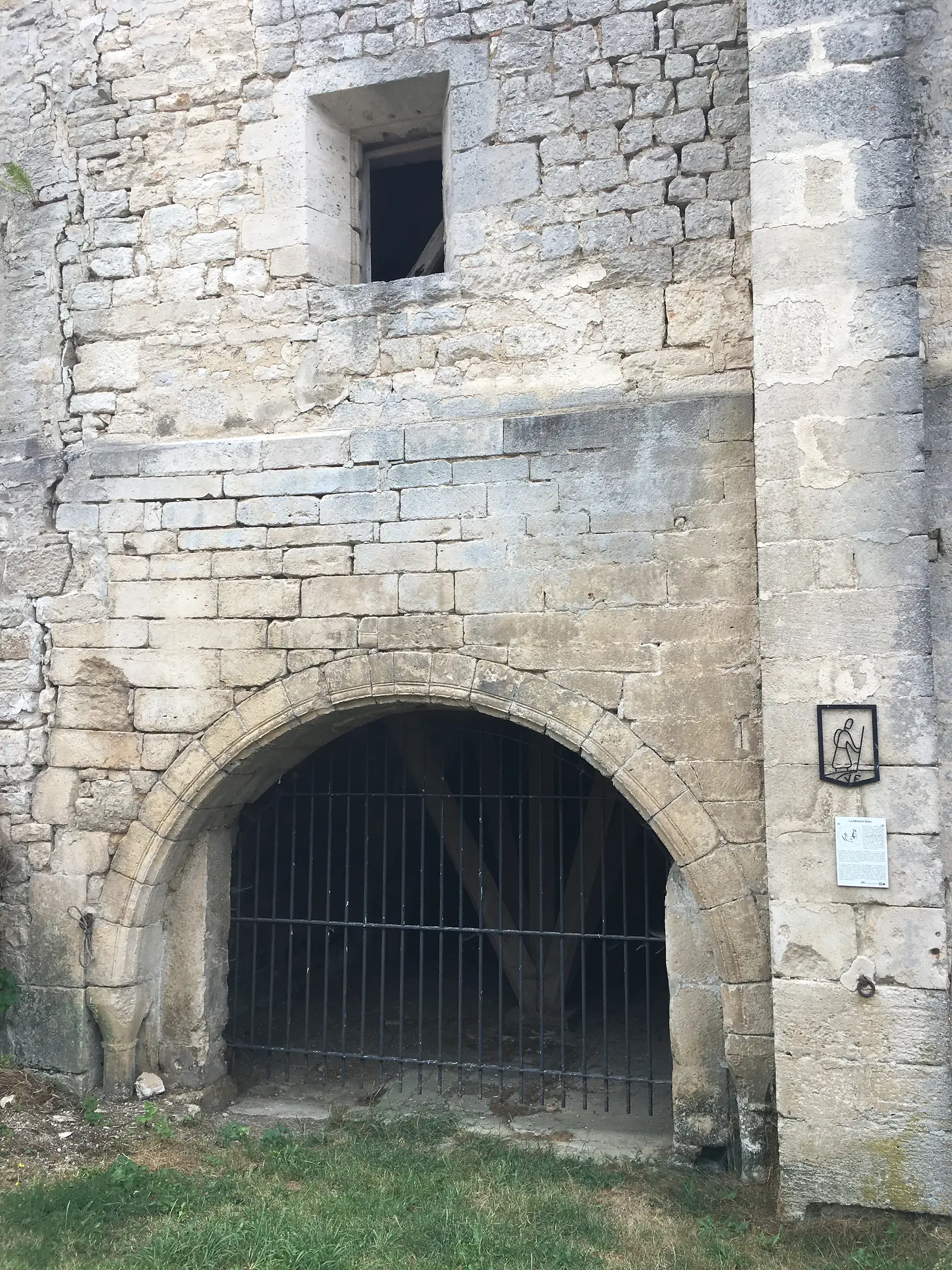 A stone culvert arch with an iron grille set into an old fortified wall, with a small barred opening alongside and grass at its base.