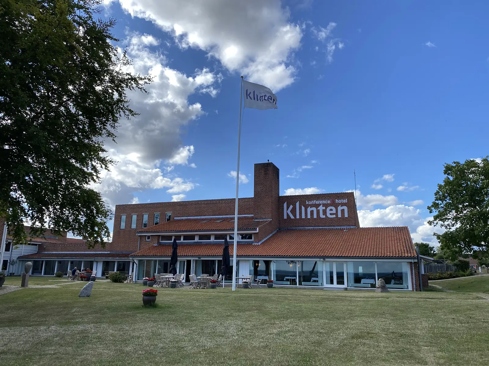 A red-brick hotel lettered "konference & hotel klinten" with a flag on a mast and cafe tables on the lawn under cumulus.