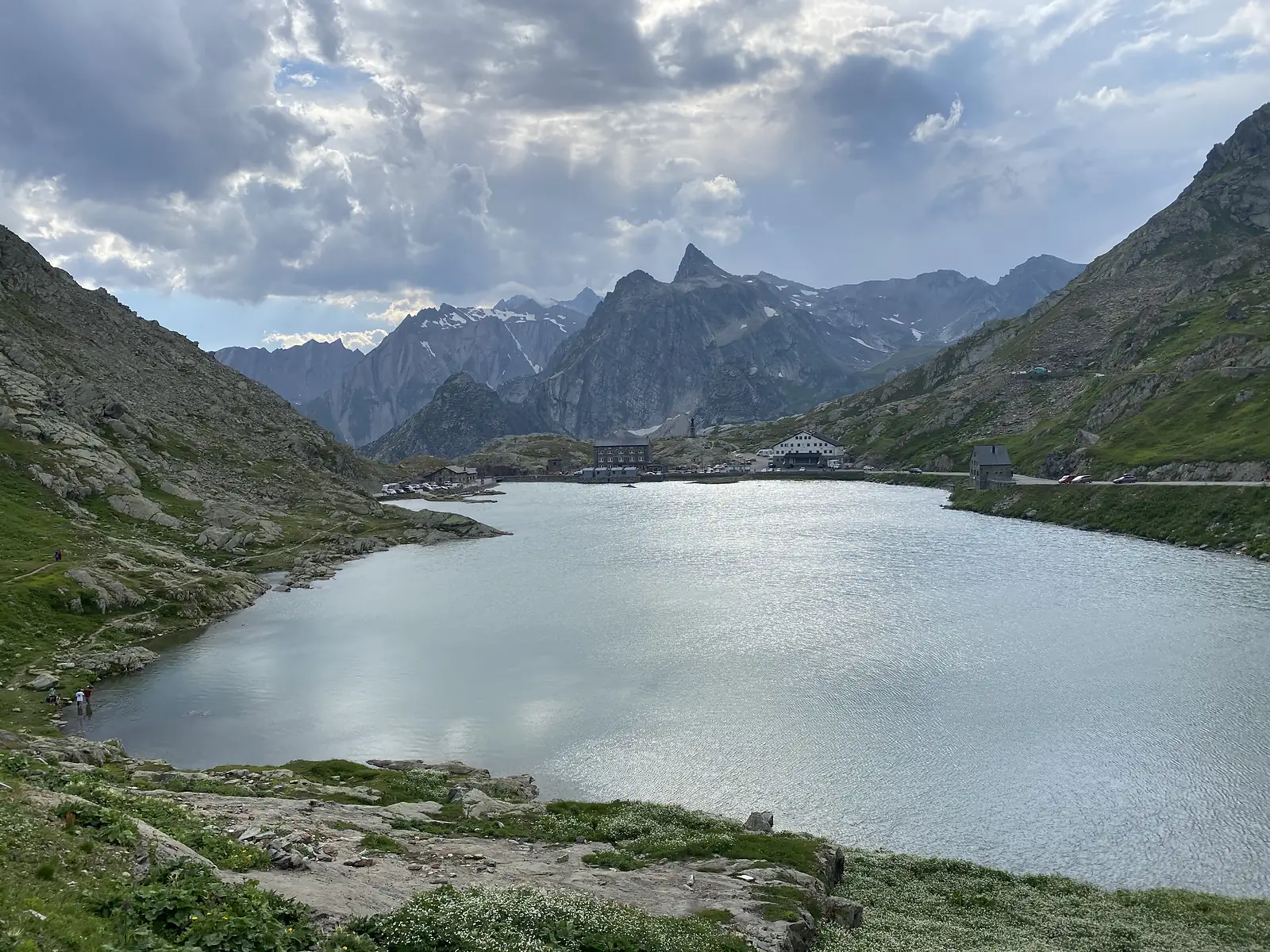 
The pass lake lit by hazy sunshine breaking through heavy cloud, with the hospice and roadside buildings strung along its edge.
