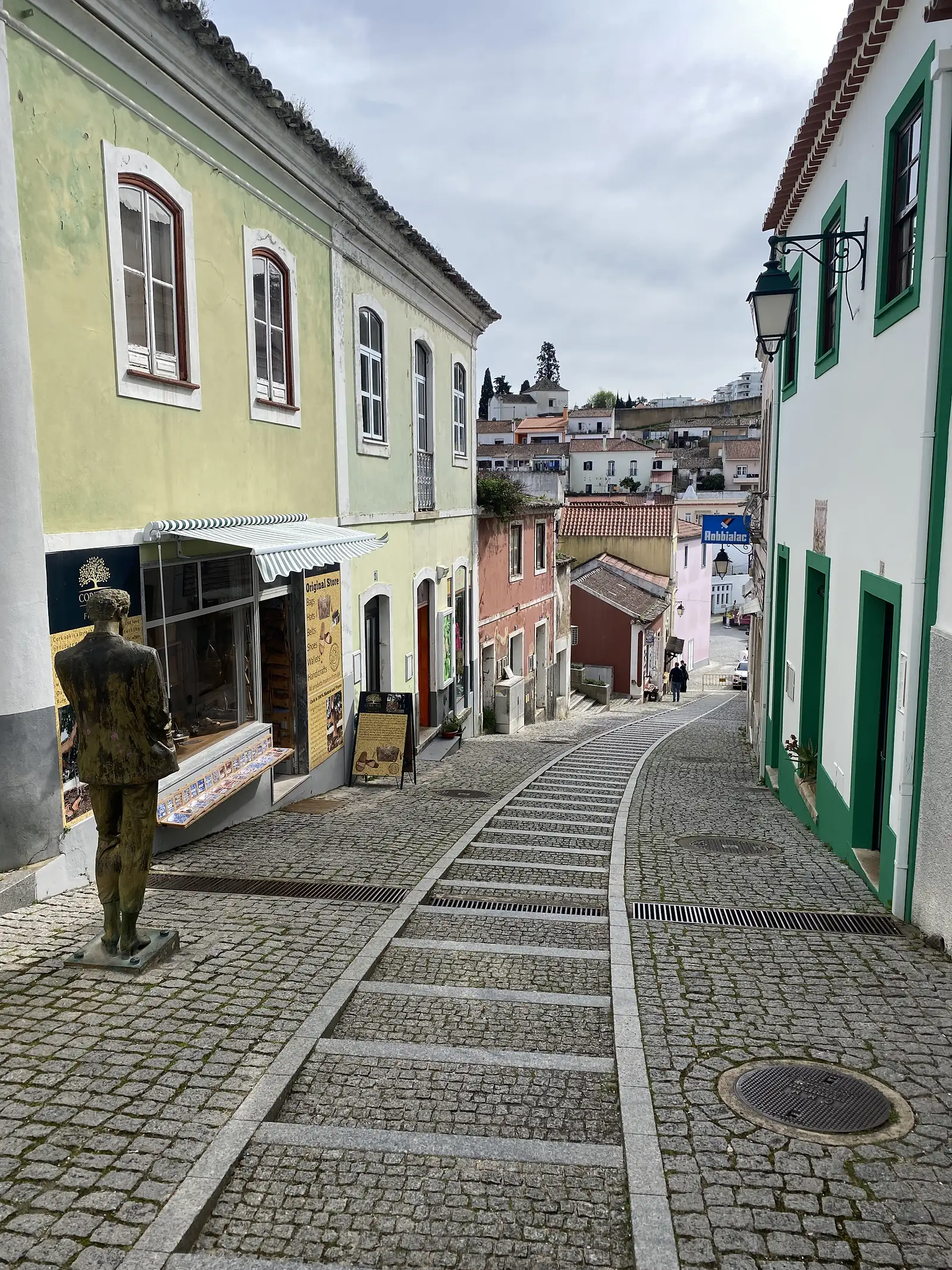 Steep cobbled town street lined with pastel-green and terracotta houses and a shopfront under a striped awning, a life-size bronze statue of a man standing on the pavement in the foreground.