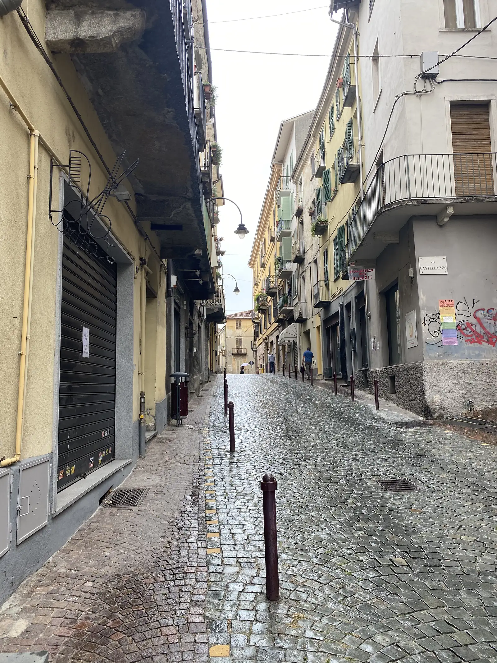 A wet cobbled street named Via Castellazzo runs between tall pastel apartment buildings with shuttered windows and wrought-iron balconies in Ivrea.