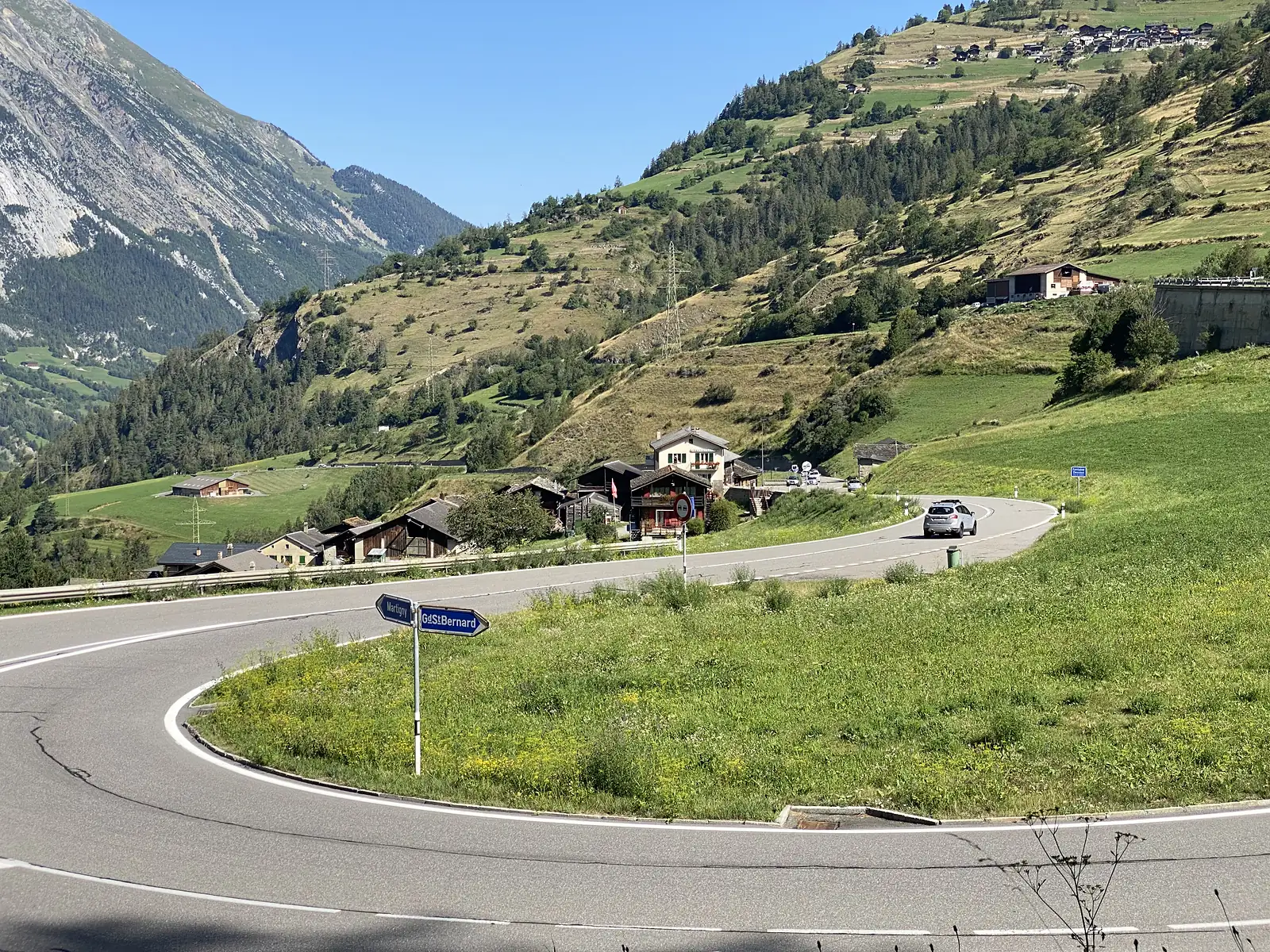 A road sign reading Martigny and GdStBernard pointing along a hairpin bend through an alpine village, with a car descending and steep green slopes above.