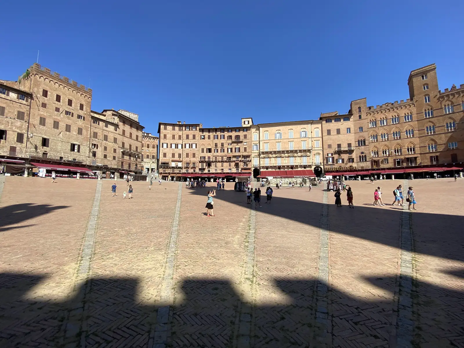 Visitors cross the fan-shaped brick paving of Siena's Piazza del Campo, ringed by tall medieval palazzi and a crenellated tower.