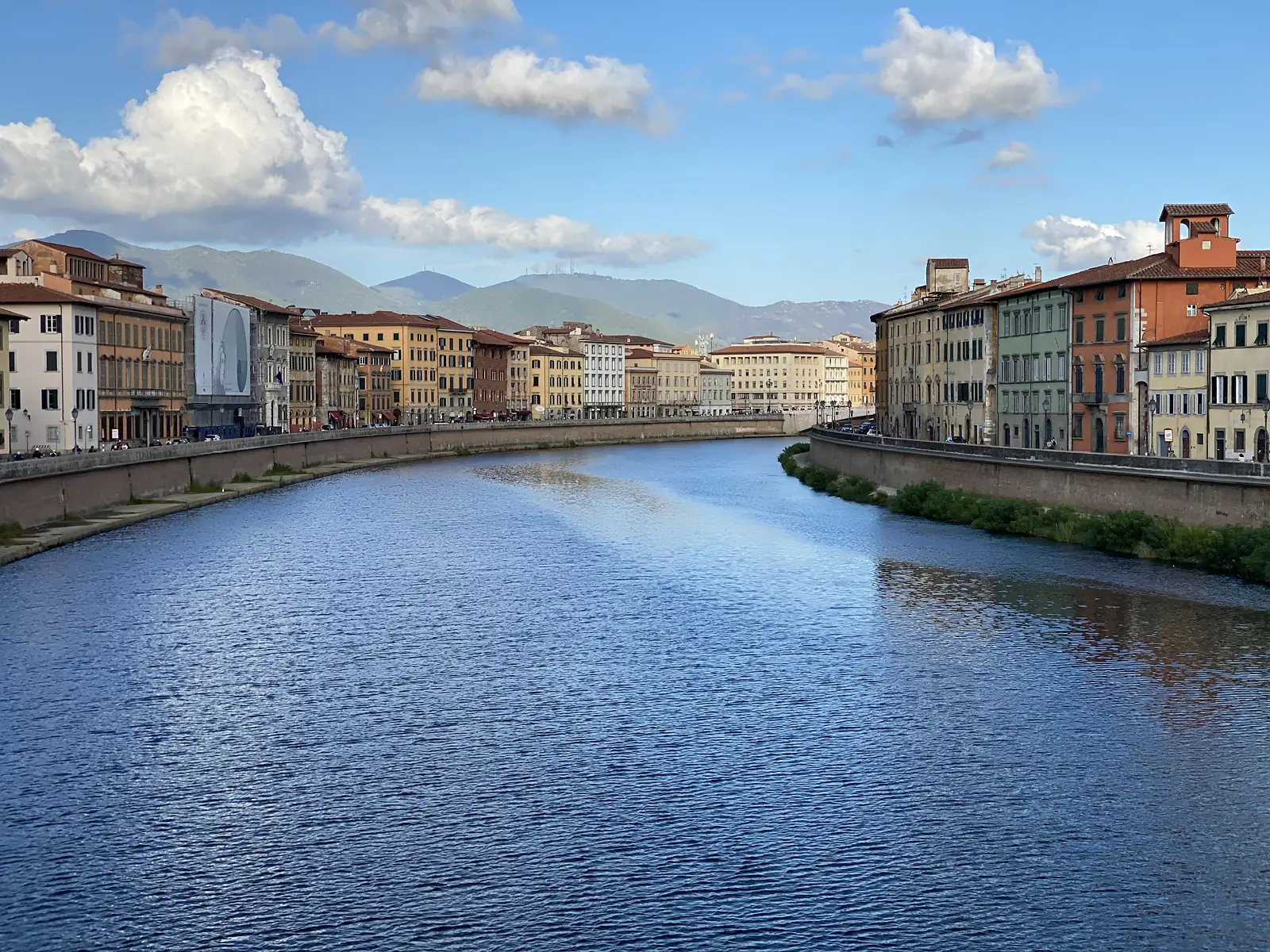 The River Arno curves past rows of pastel-coloured townhouses in Pisa, with wooded hills rising beyond the rooftops.