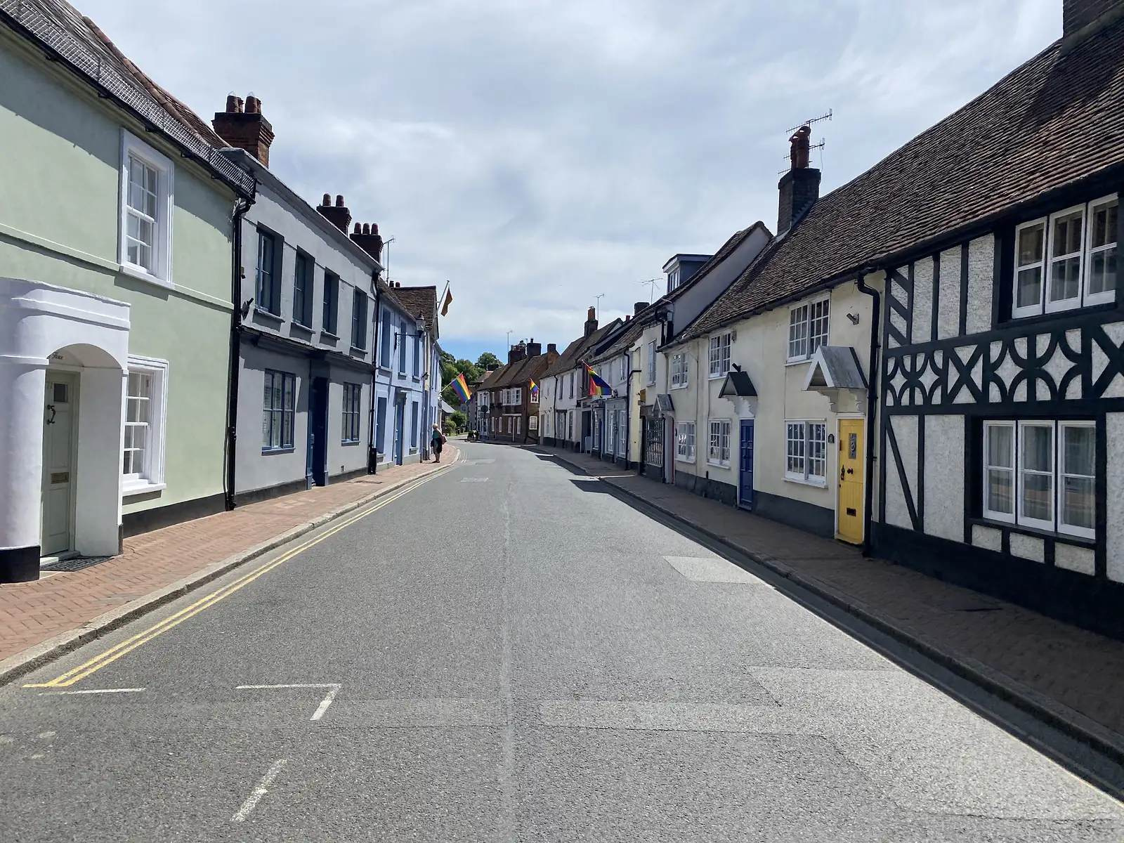 An empty small-town street of pastel-painted Georgian houses and black-and-white timbered buildings, with rainbow flags hung out.