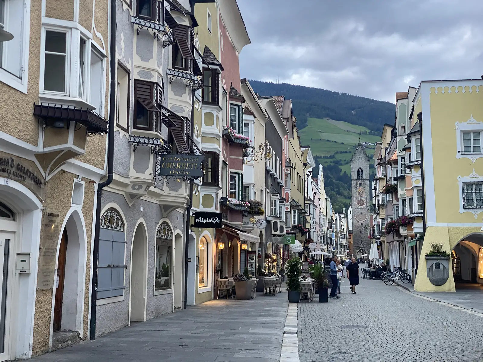 An old-town street of painted, bay-windowed houses and arcades runs down to a tall stone clock tower, with a green hillside rising behind under a grey sky.