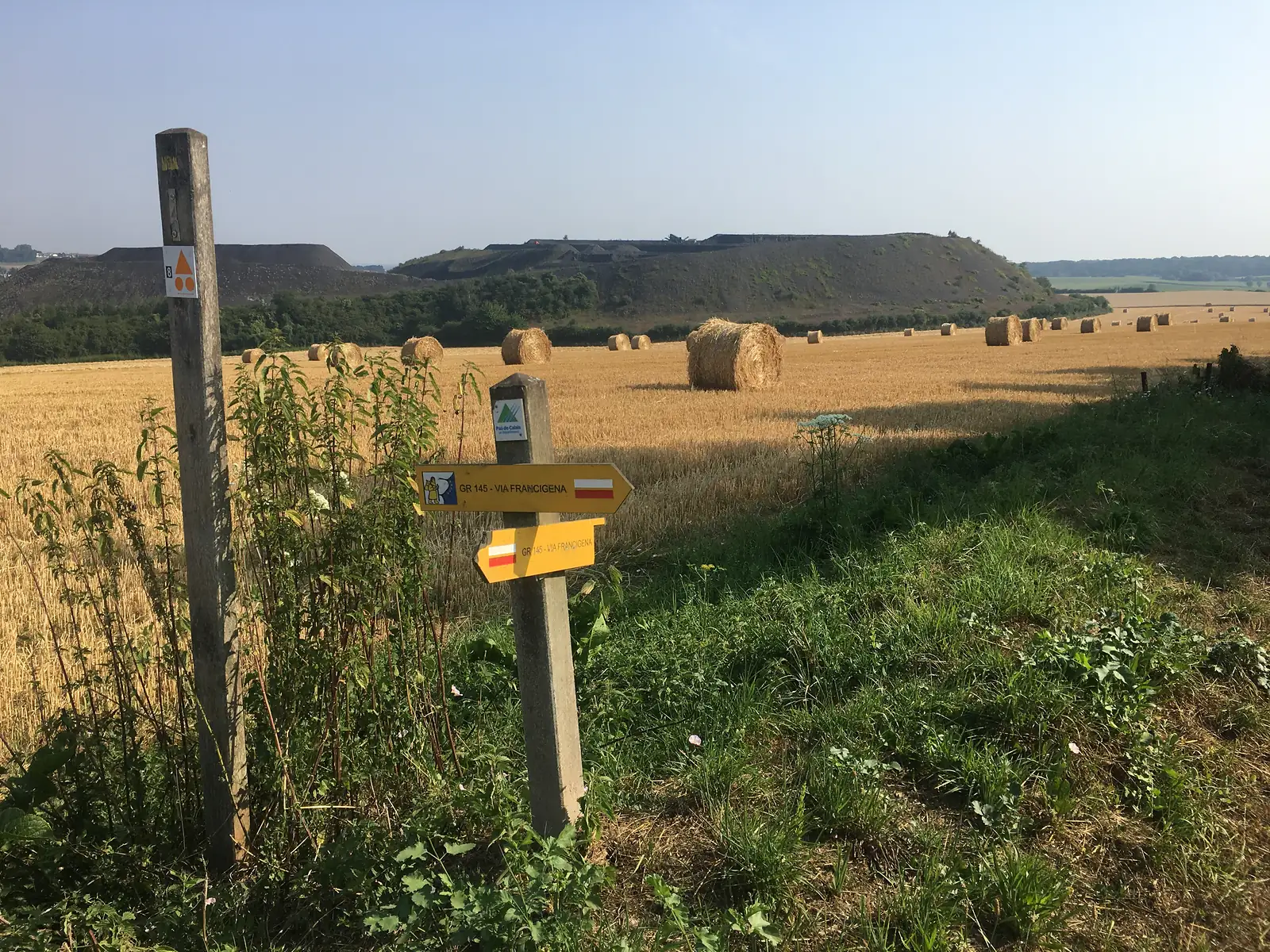 A waymarker post reading 'GR 145 - Via Francigena' pointing across a stubble field scattered with round straw bales, a spoil heap from a former coal mine rising in the distance.