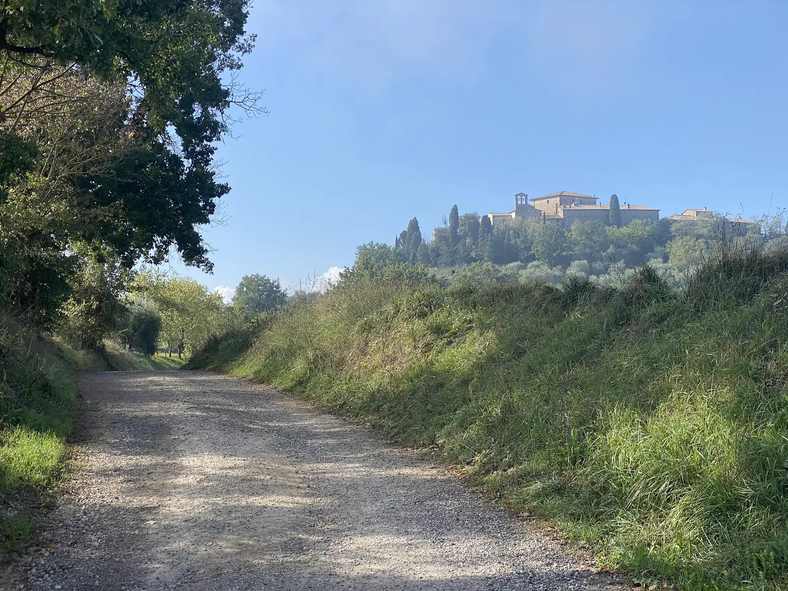 A gravel road climbing towards a stone hilltop hamlet with a bell tower, seen across grassy banks under a clear sky.