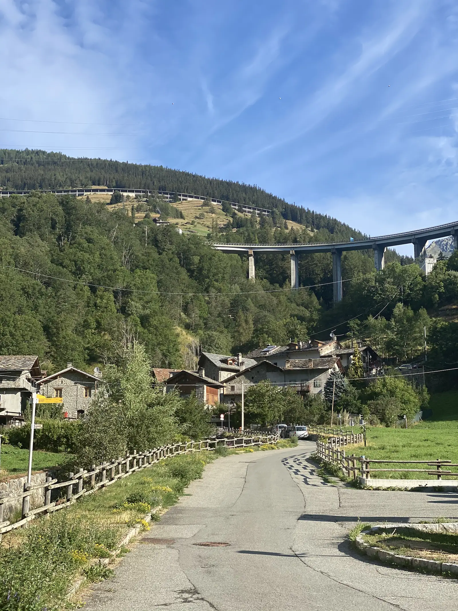 A tall grey road viaduct spans a forested valley above a small stone village, a paved lane leading down past wooden fences toward the houses.