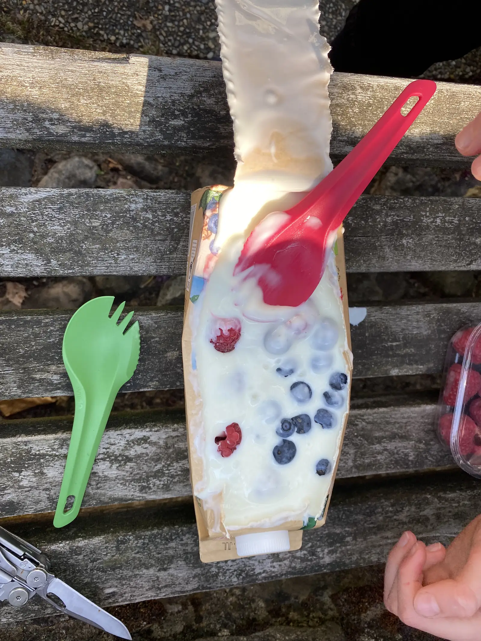 An improvised breakfast on a weathered bench: a slit-open milk carton filled with yoghurt, blueberries and raspberries, with a green spork and a multi-tool alongside.