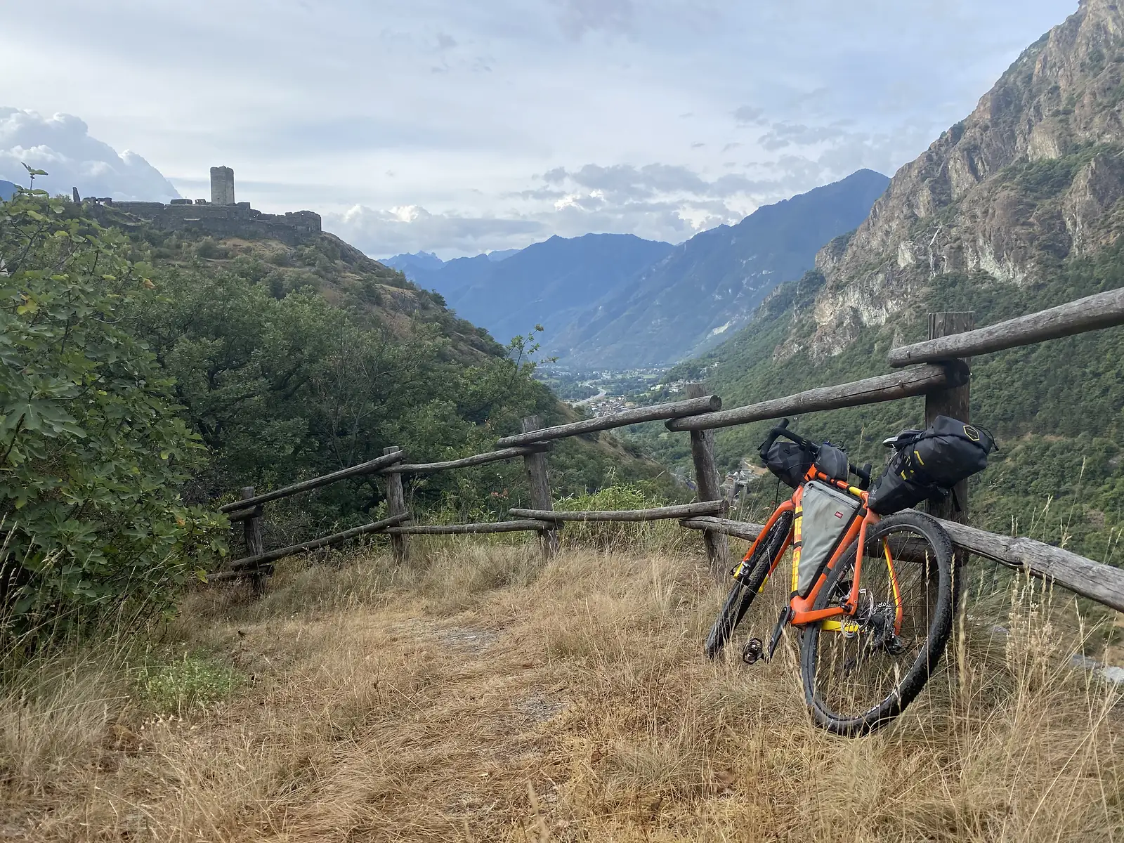 A loaded orange gravel bike leaning against a wooden fence at a viewpoint, with the ruined tower of a hilltop castle and a valley town visible in the distance.