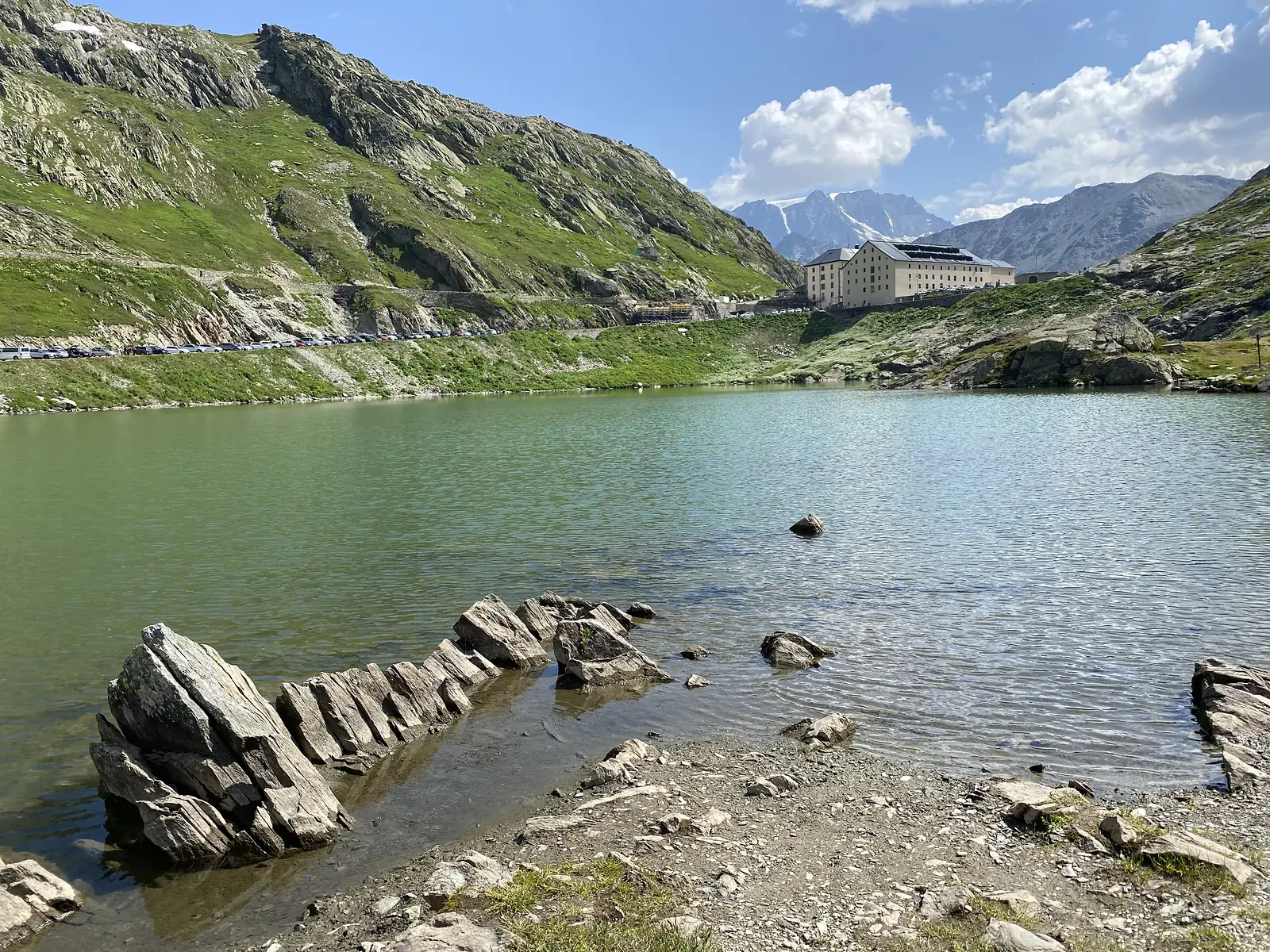 A stone breakwater of stacked slabs at the lake's edge, with the hospice building reflected across the calm green water.