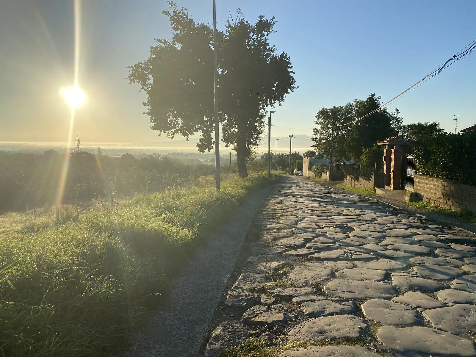An old cobbled lane running along a ridge past gated villas, low sun flaring above misty countryside on the other side.