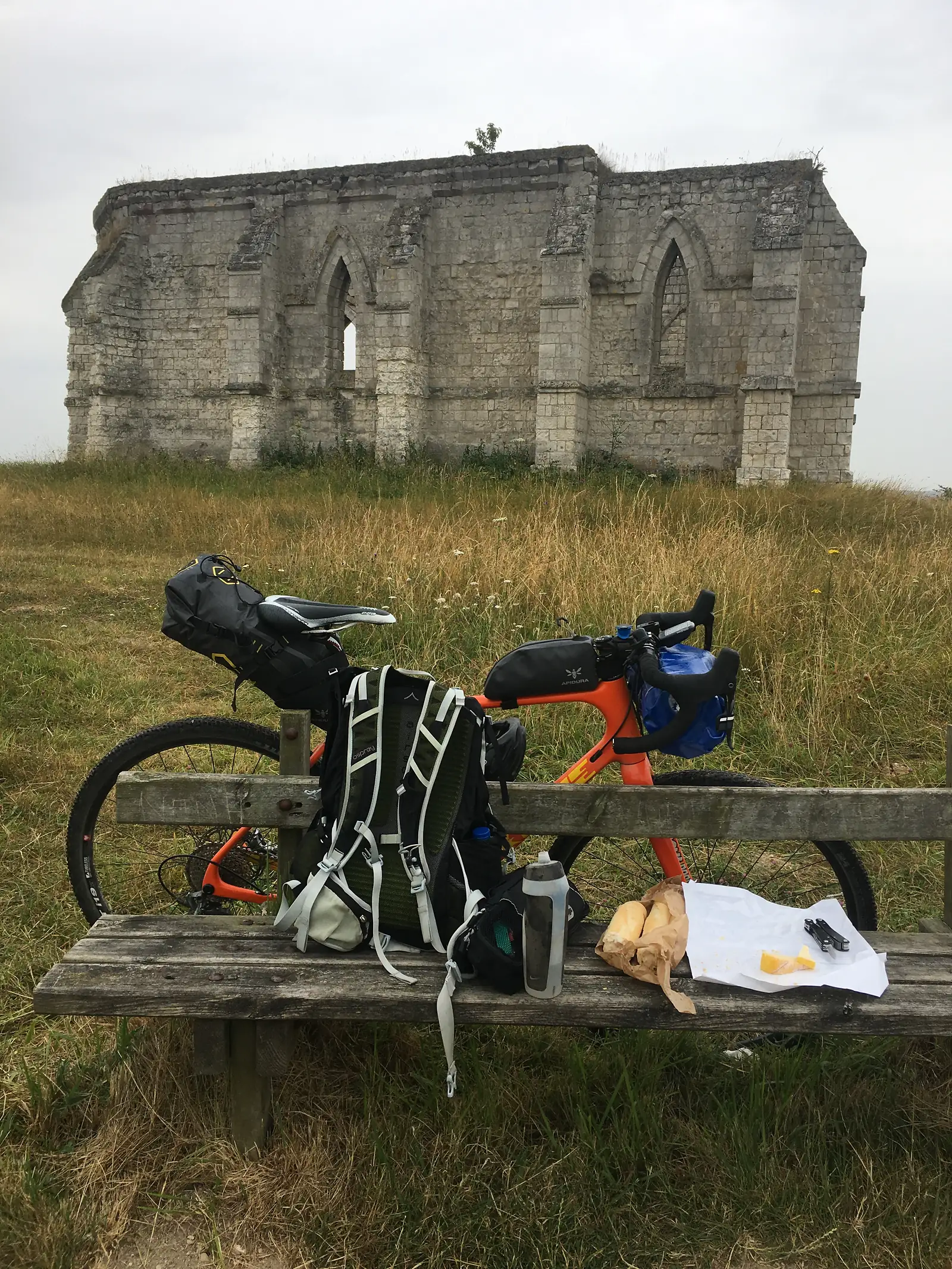 A loaded orange gravel bike rests against a wooden fence rail with a baguette and wrapped food propped beside it, the ruined chapel visible in the background.