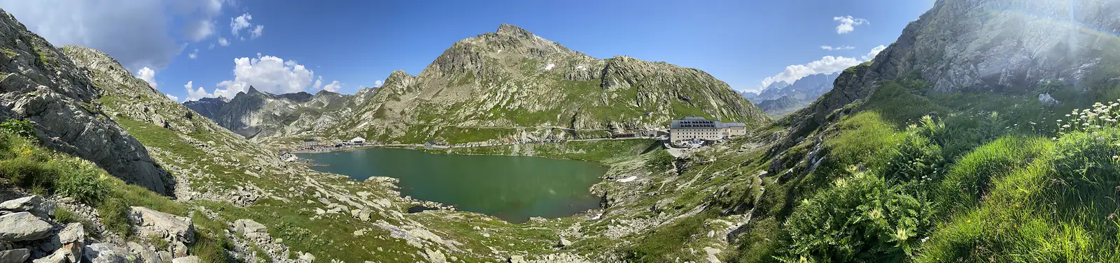 A wide panorama of the green pass lake ringed by rocky peaks, with the hospice buildings and the road curving along its northern shore.