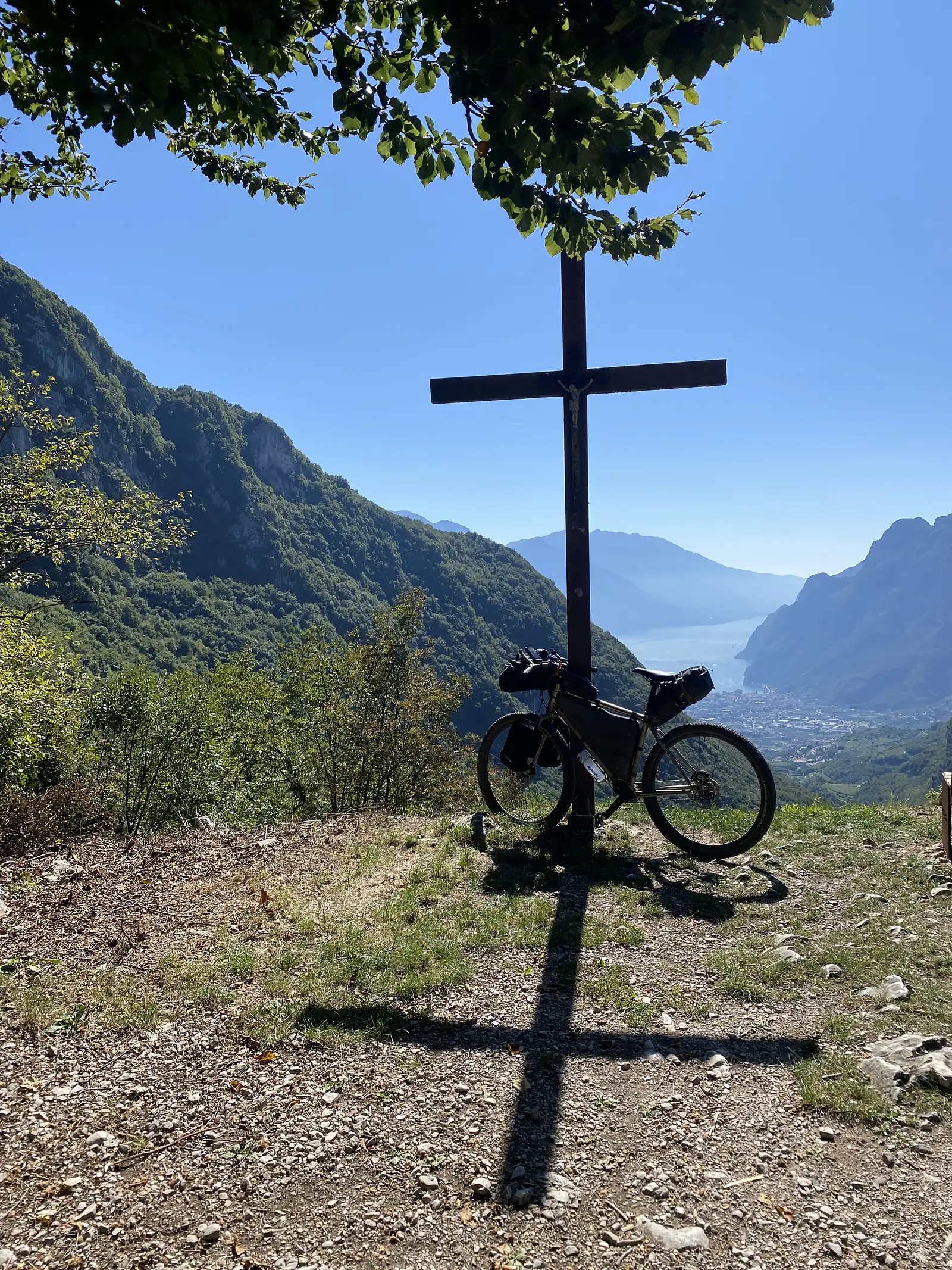 A loaded bikepacking bike leans against a tall iron crucifix on a grassy viewpoint, with a lake and town far below between steep mountain walls.
