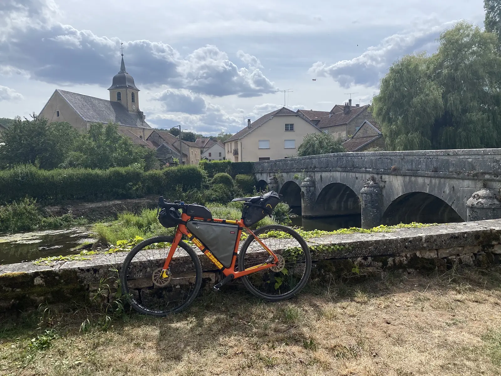 A loaded orange gravel bike leans against the parapet of a multi-arched stone bridge over a river, a village with a church spire and a weeping willow behind.