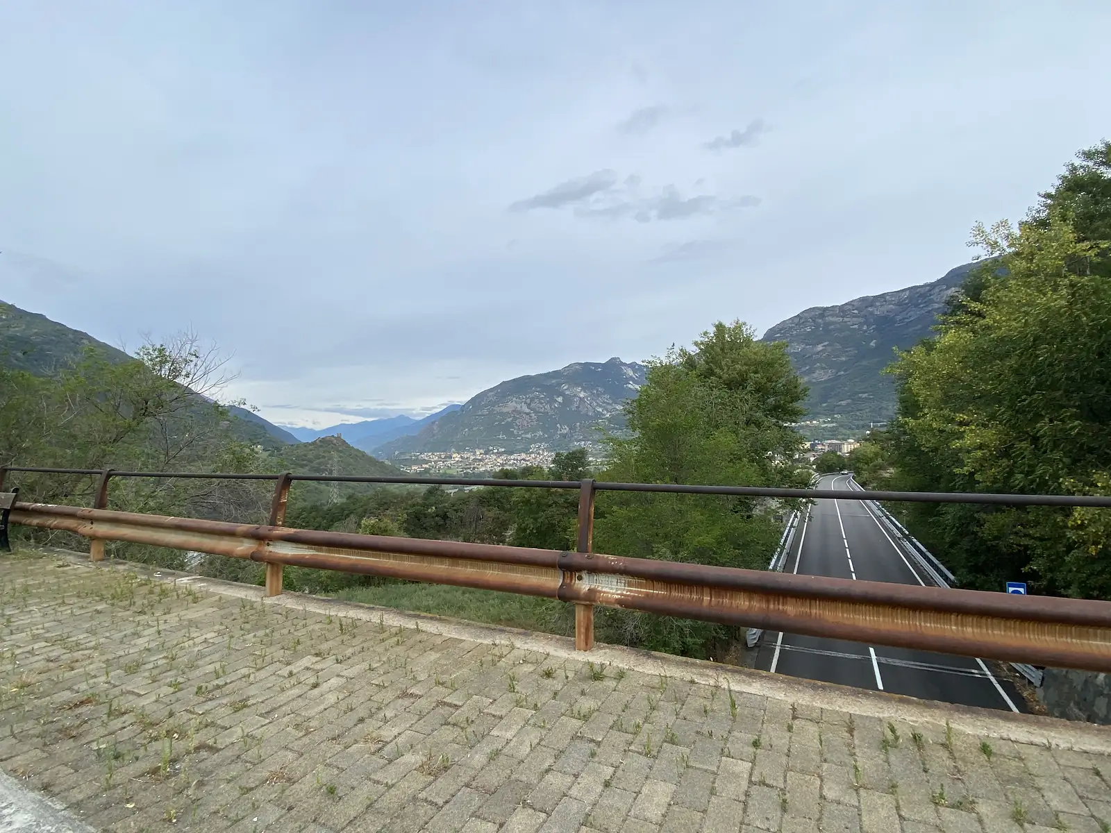 A rusted crash barrier on a hillside viewpoint overlooking a road that curves down through the Aosta Valley towards a hazy town below.