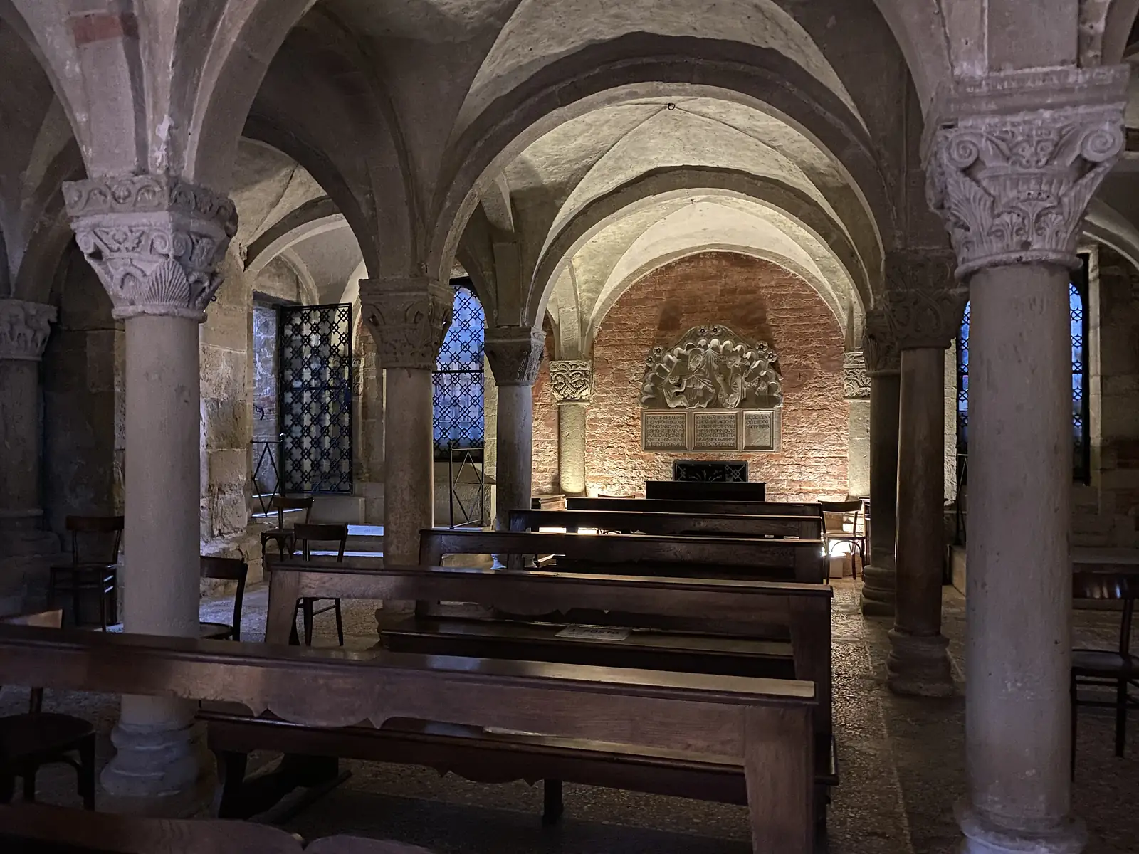 Wooden pews sit beneath the vaulted, carved capitals of a Romanesque crypt, lit warmly against a brick apse decorated with a carved relief.