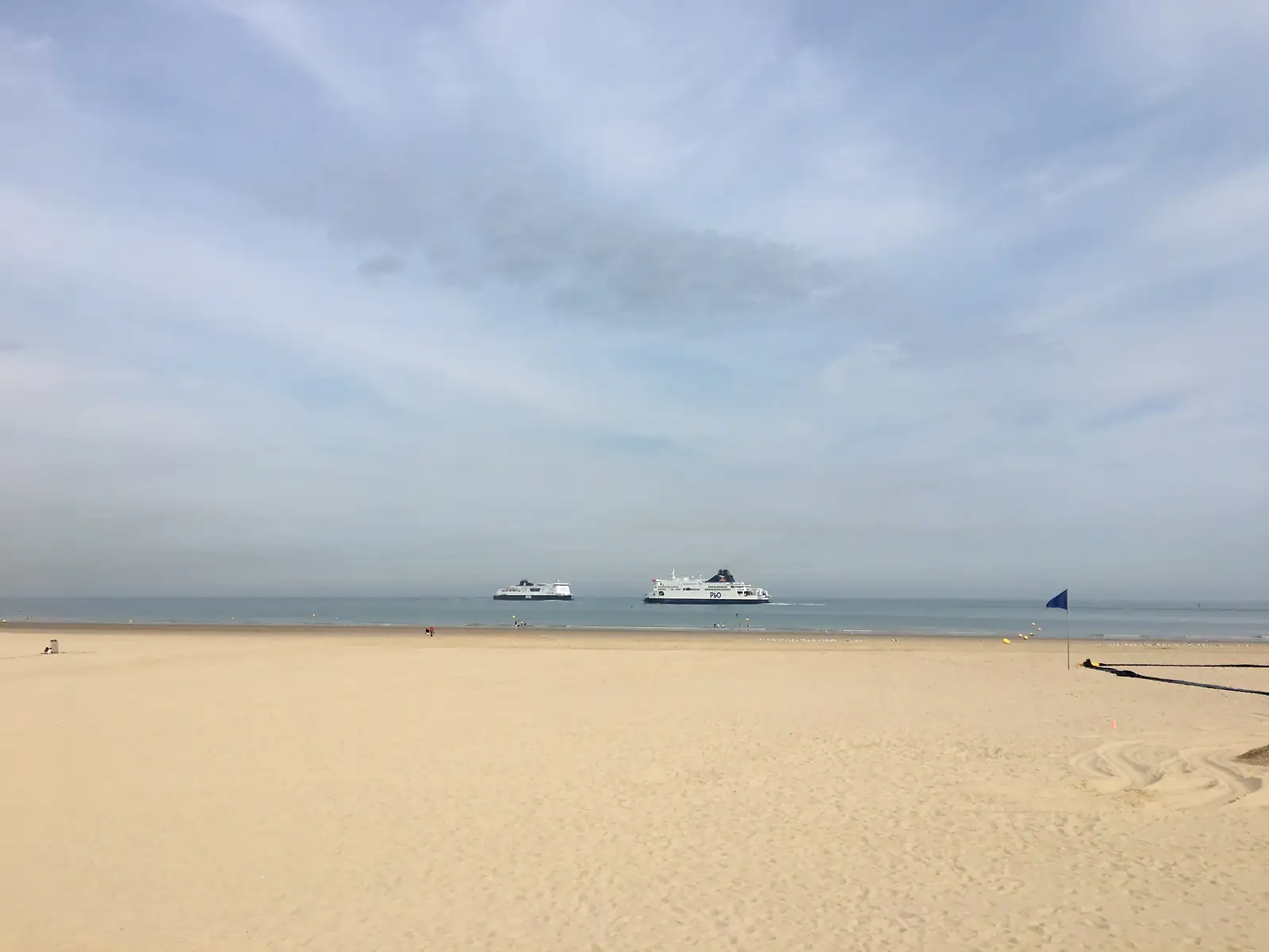 Two cross-Channel ferries, one marked P&O, passing each other at sea beyond an empty sandy beach with a blue flag flying near the shoreline.