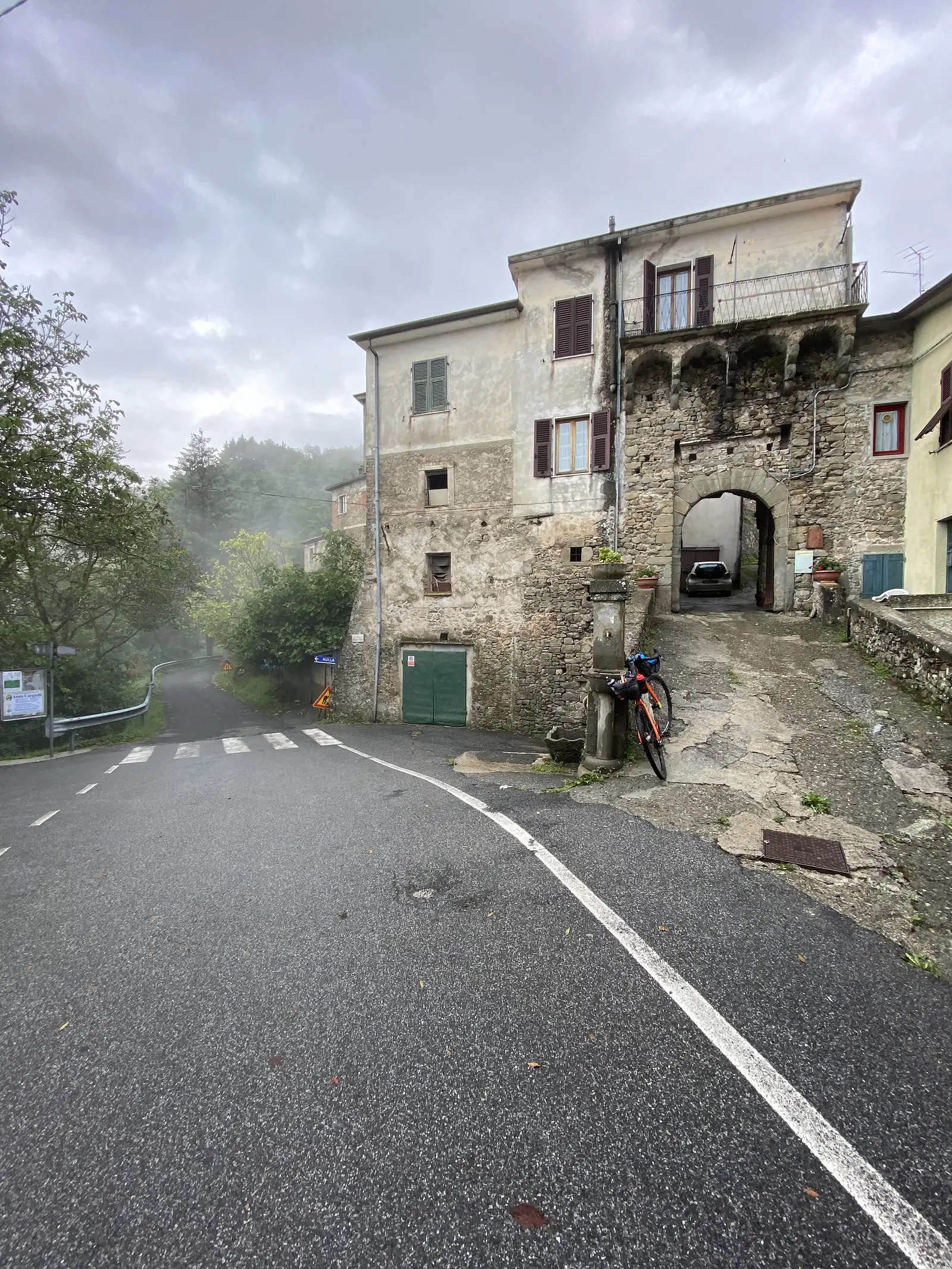 A public stone water trough and tap stand beside old stone houses on a village street, with a loaded touring bike parked against it and a blue "Bulla" road sign pointing the way.