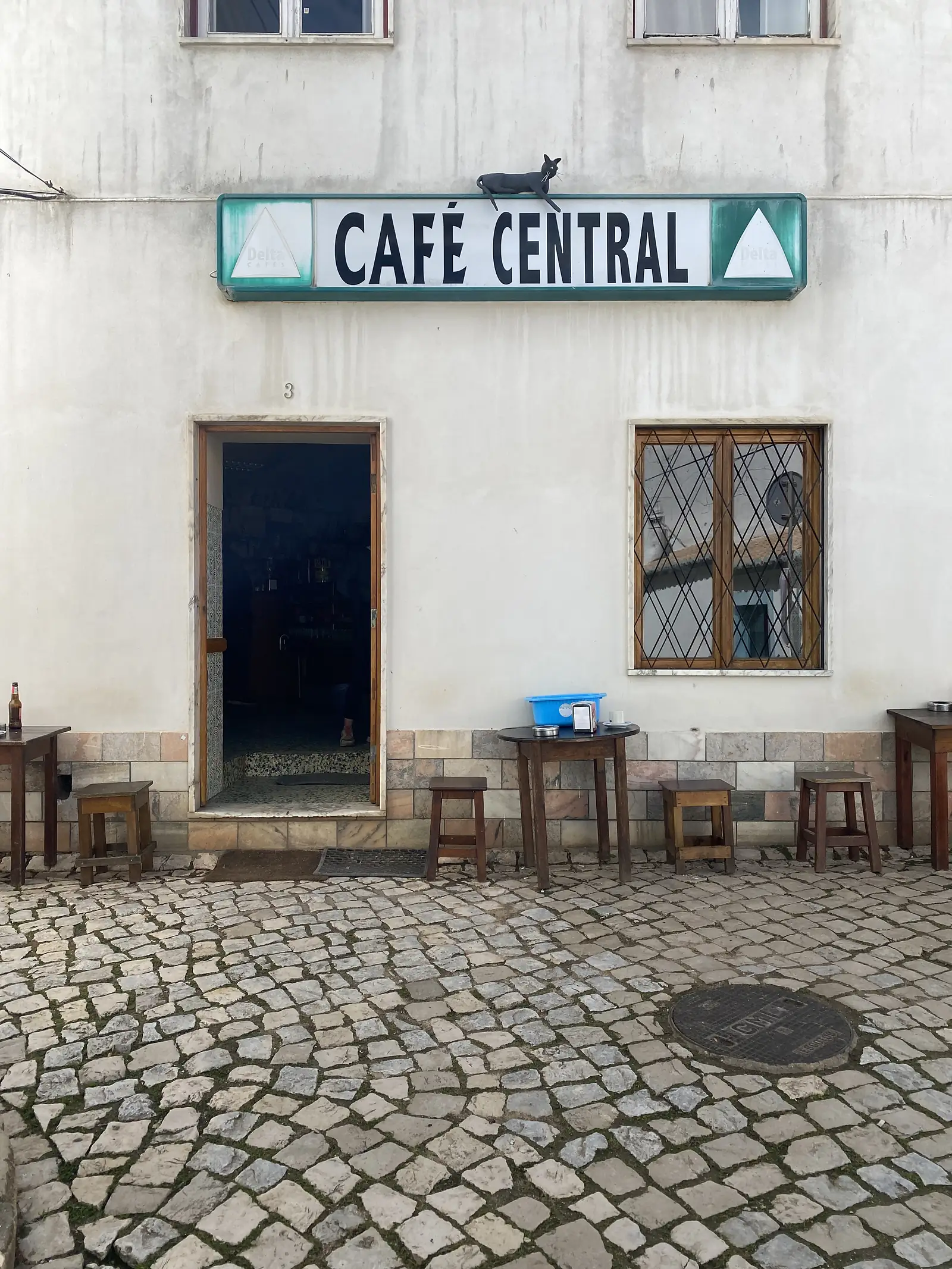 Whitewashed café frontage with a hand-painted "CAFÉ CENTRAL" sign, a black cat sitting on top of it, and wooden stools and small tables set out on the cobbles beside an open doorway.
