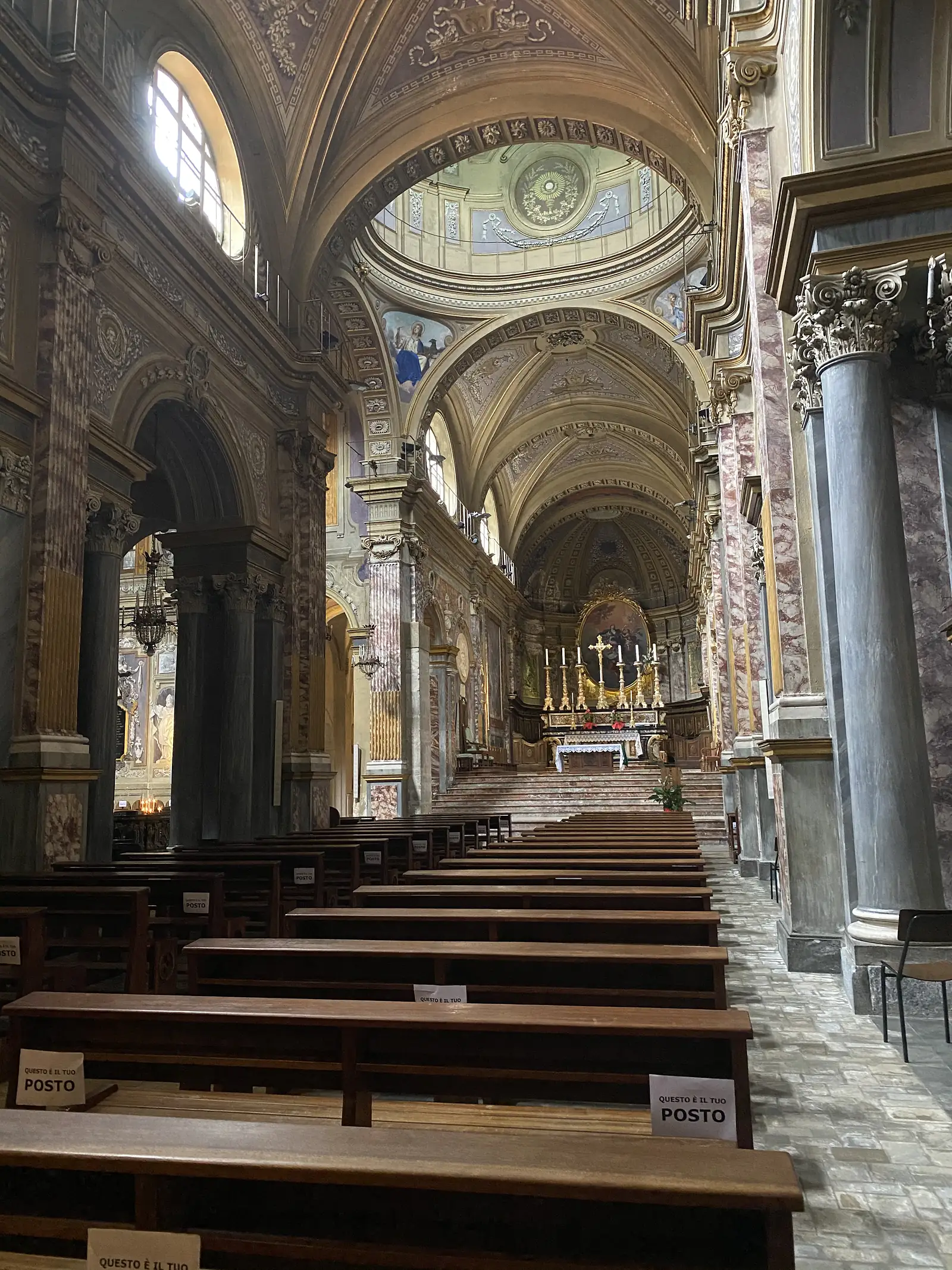 The nave of a richly decorated Baroque church looking towards a gilded high altar beneath a painted dome, its wooden pews carrying paper signs reading "Questo è il tuo posto".