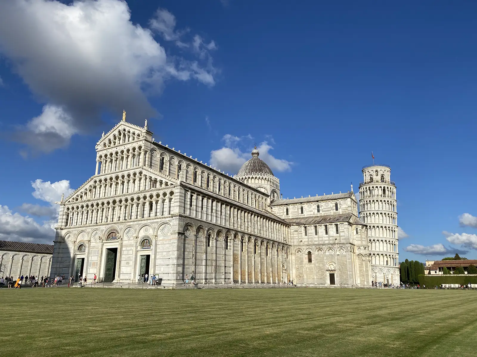 Pisa Cathedral's long marble flank and dome stand beside the Leaning Tower across a wide green lawn, tourists strolling in the foreground.
